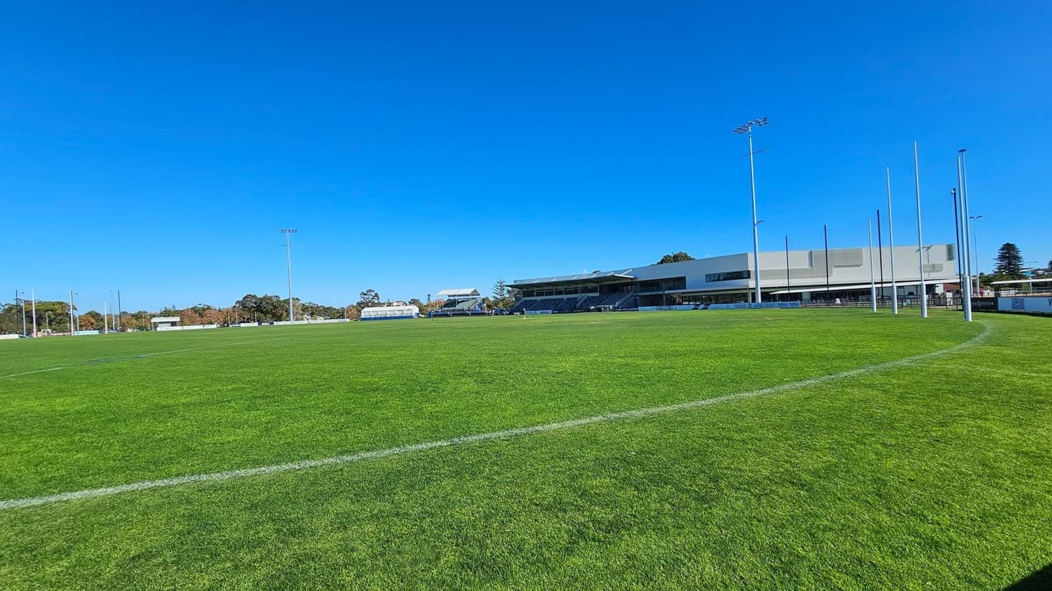 A large, well-maintained grassy sports field with white boundary lines. On the right, there are several tall goalposts and a modern building with a flat roof and large windows. In the background, there are more structures, including a smaller building and some trees. The field is surrounded by a low fence, and there are tall floodlights around the perimeter. The sky is clear and blue.