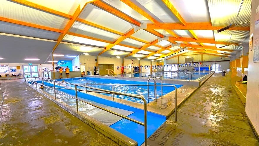 Side view of the low-set building of Huntly Aquatic Centre with blue and yellow accents. The large font of “Huntly Aquatic Center” is clearly detailed on the front wall of the facility. There is a red, teardrop-shaped flag that says, “Pool Open” just before the entryway on the side that has steel handrails. The building is close to a parking space and an electric utility pole and is surrounded by tall trees.