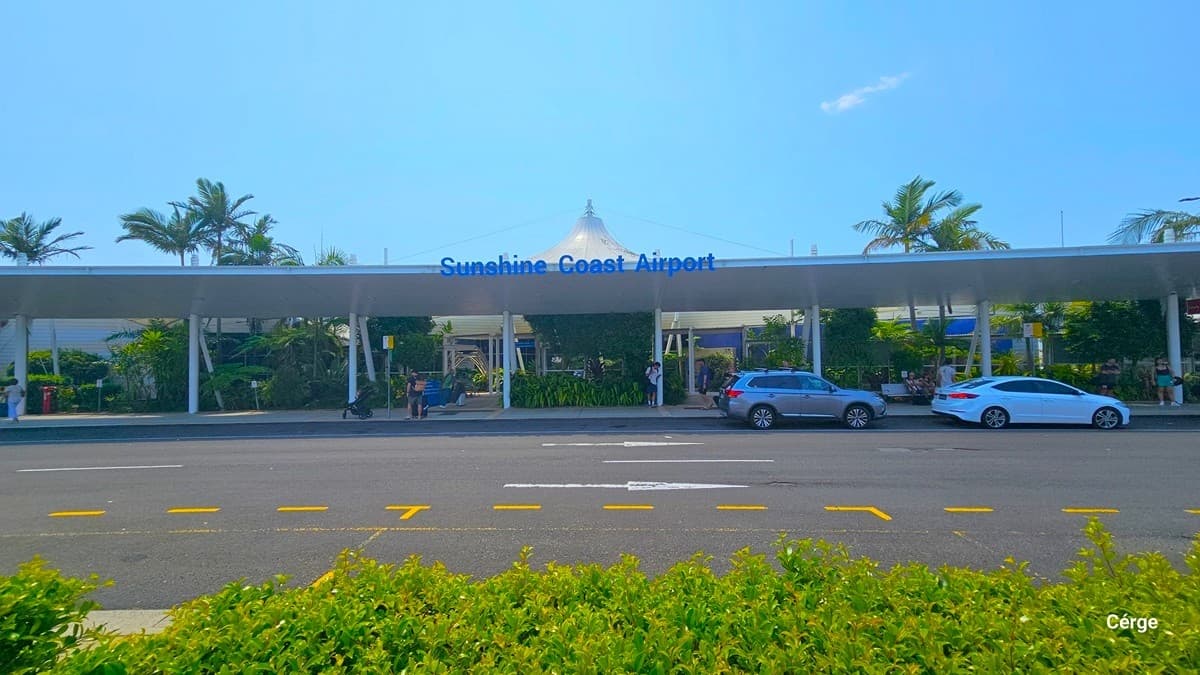 A covered airport departures area with a long, white canopy extending across the scene. On the left, there are construction barriers and signs, including a blue "Departures" sign. In the middle, a pedestrian crossing with red and white stripes leads to the road. On the right, a red vehicle is parked near the kerb. Palm trees are visible in the background, and the road has markings for passenger drop-off. The area is well-lit with overhead lights.