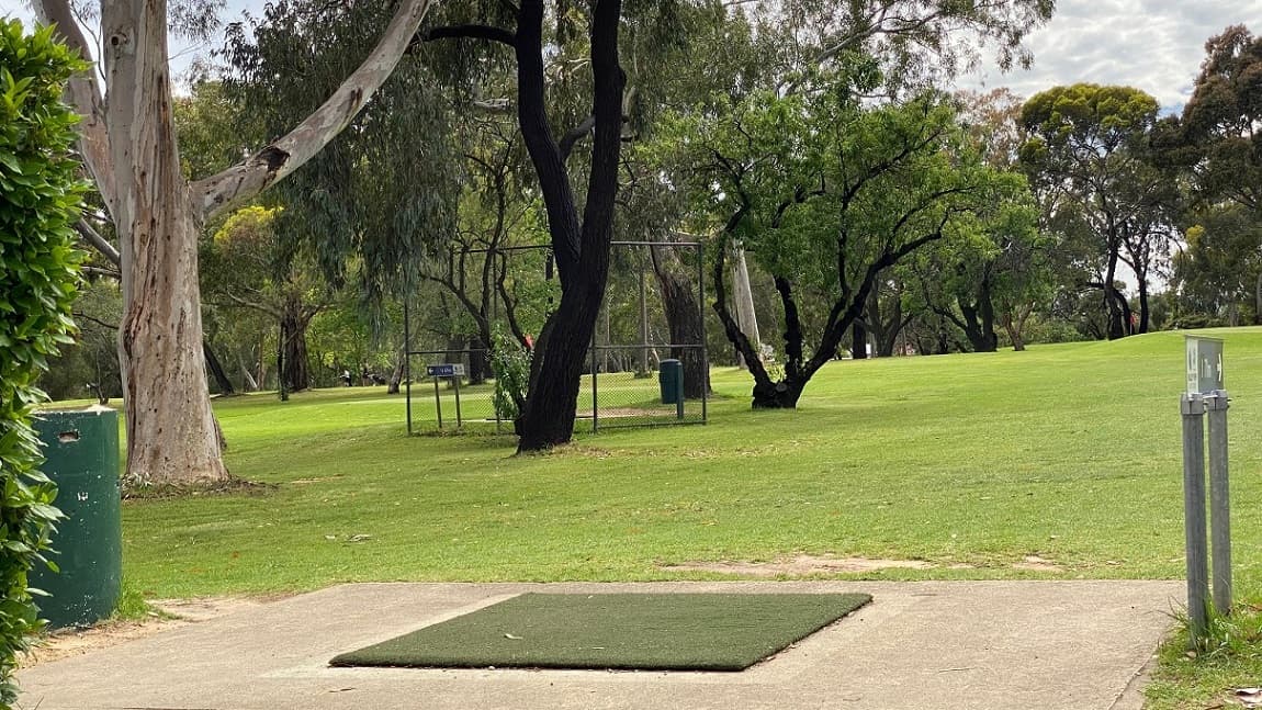 A concrete path features a square of artificial grass with a small, green structure to the left. To the right is the notice board providing direction to the golf course ahead. has rail on both sides and overlooks a golf course. A golf course fence is in the centre with a direction board and a green-painted pillar.