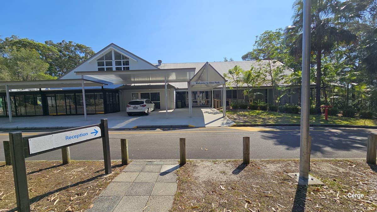 A paved area with a circular garden bed in the centre, surrounded by a road. Sparse trees and shrubbery populate the garden bed and the periphery. Behind is a building with a large triangular roof feature and an extended wing. Tall green trees frame the background.