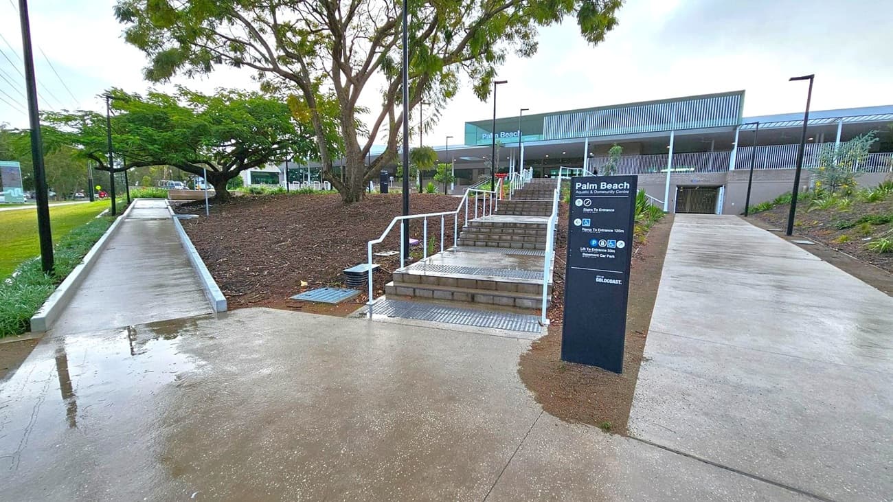 A concrete pathway leads to a set of stairs with white railings in the centre, flanked by a sign for Palm Beach Aquatic & Community Centre. To the left, a sloped path runs alongside a grassy area with a large tree. On the right, another path leads to the building entrance. The building in the background has a modern design with a large overhang and louvred facade. The ground is wet, suggesting recent rain, and there are street lamps along the paths.