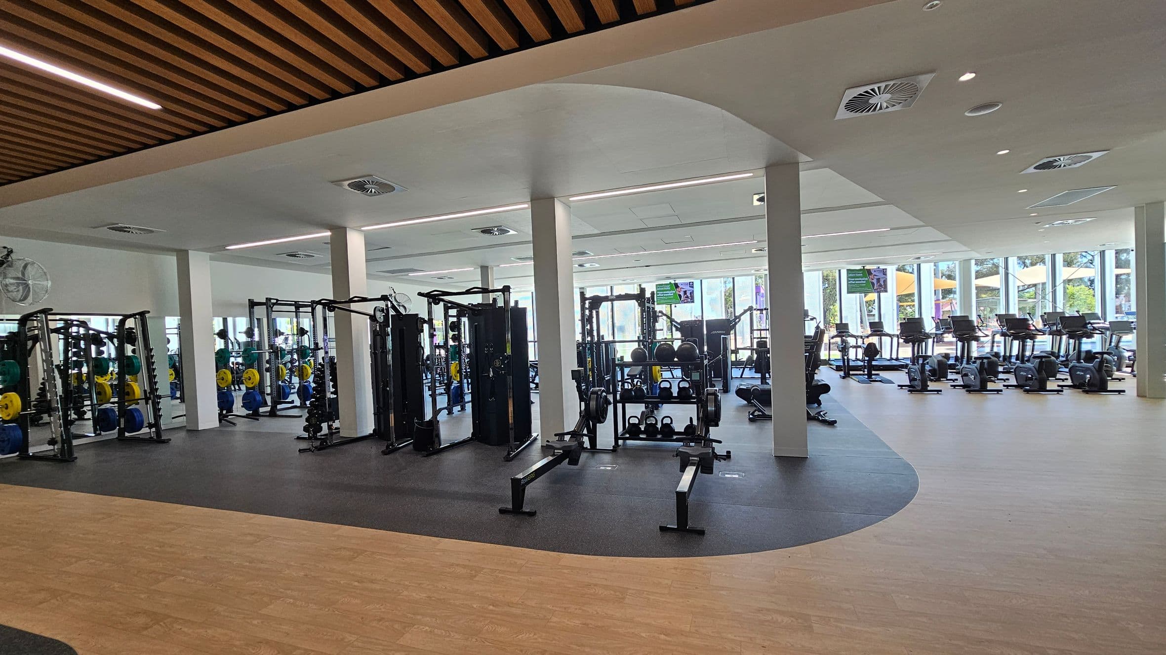 Glass doors open into a health club. Inside, the floor is light wood, and the ceiling has wooden slats. On the left, there are exercise mats and weights against a white wall. In the centre, gym equipment is visible, including weight machines and racks. The right side has a striped wall and a sign for hearing assistance. The entrance has a grey floor and a sign with "ACTIVE" branding.