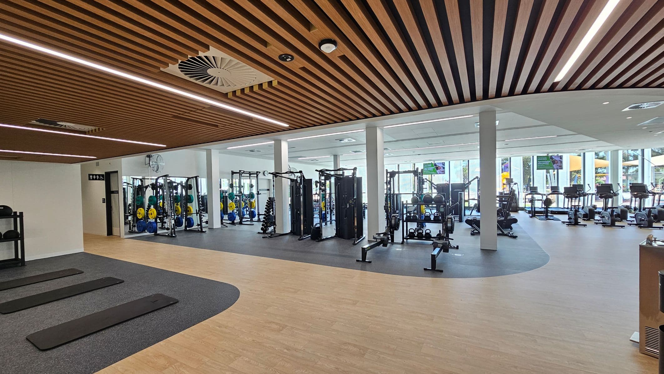 A modern gym interior with a wooden slatted ceiling and recessed lighting. On the left, there are exercise mats on a grey floor section. In the centre, weightlifting equipment and racks with colourful weights are visible. The right side features cardio machines like treadmills and rowing machines near large windows. The floor transitions from wood to a darker material in the equipment area. White pillars support the ceiling, and a wall-mounted fan is visible.