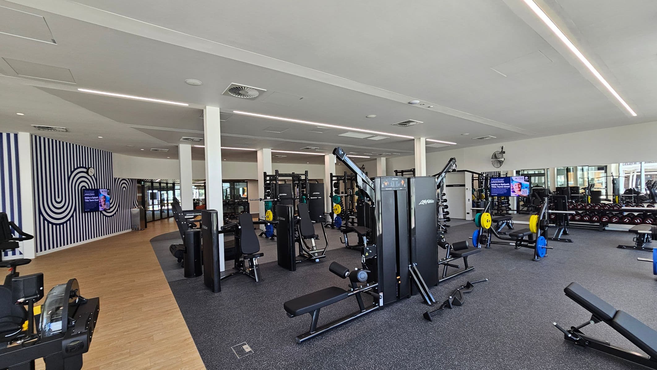 A spacious gym with various exercise equipment. On the left, there's a wall with a striped pattern and a digital screen. The floor transitions from wood to grey carpet. In the centre, there are weight machines and benches. On the right, a mirrored wall reflects more equipment, including weight racks and benches with colourful weight plates. The ceiling is white with recessed lighting and air vents.