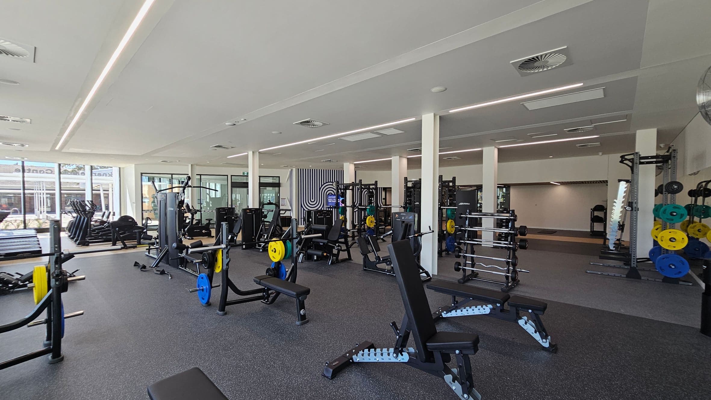 A spacious gym with various exercise equipment. On the left, there are treadmills and elliptical machines near large windows. In the centre, weight benches and machines with colourful weight plates are arranged. On the right, there are squat racks and more weightlifting equipment. The floor is covered with dark rubber mats, and the ceiling has recessed lighting and air vents.