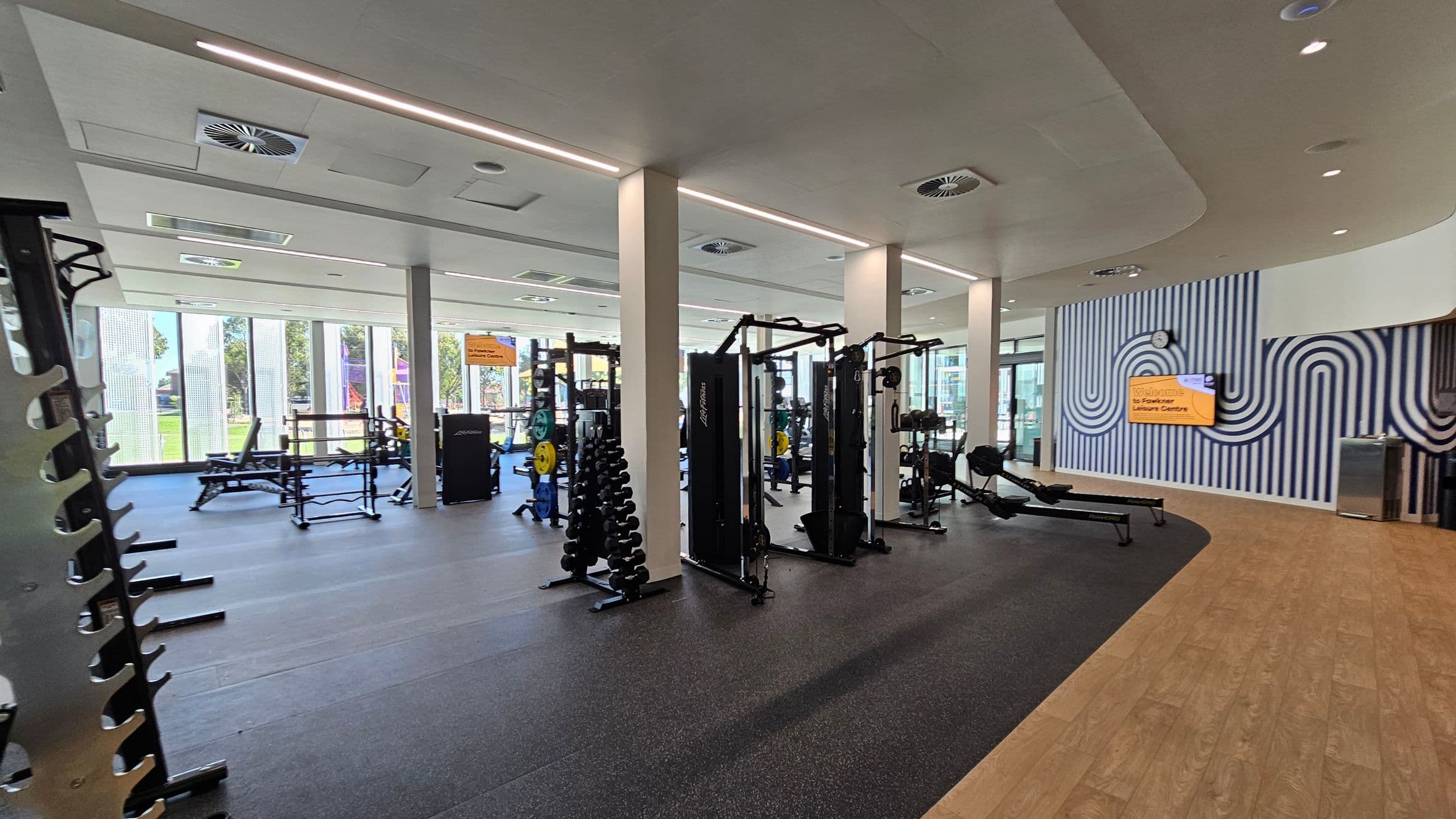 A modern gym interior with various exercise equipment. On the left, there are weight racks and benches. In the centre, there are weight machines and dumbbells. The right side features a wall with a striped pattern and a digital sign. The floor transitions from rubber matting to wood, and the ceiling has recessed lighting and air vents. Large windows in the background allow natural light to enter, with a view of greenery outside.