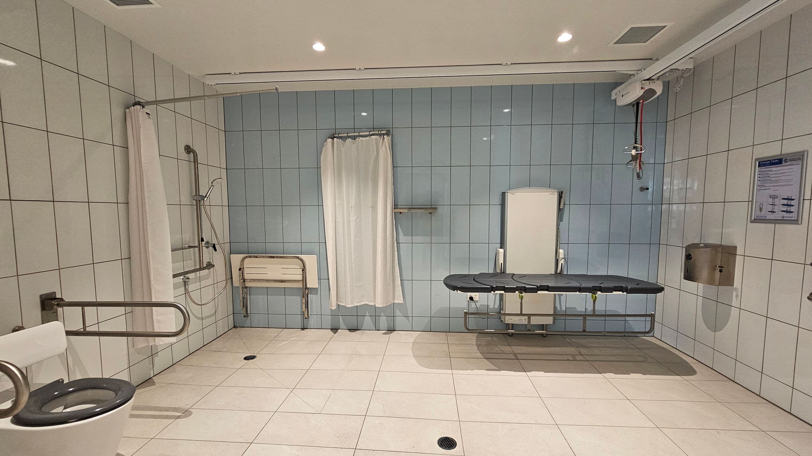 A spacious, accessible bathroom with white tiled walls and floor. On the left, there's a toilet with a blue seat and a metal grab bar. A shower area with a curtain and a foldable seat is nearby. In the centre, a blue changing table is mounted on the wall. On the right, a wall-mounted paper dispenser and a sign are visible. The ceiling has recessed lighting.