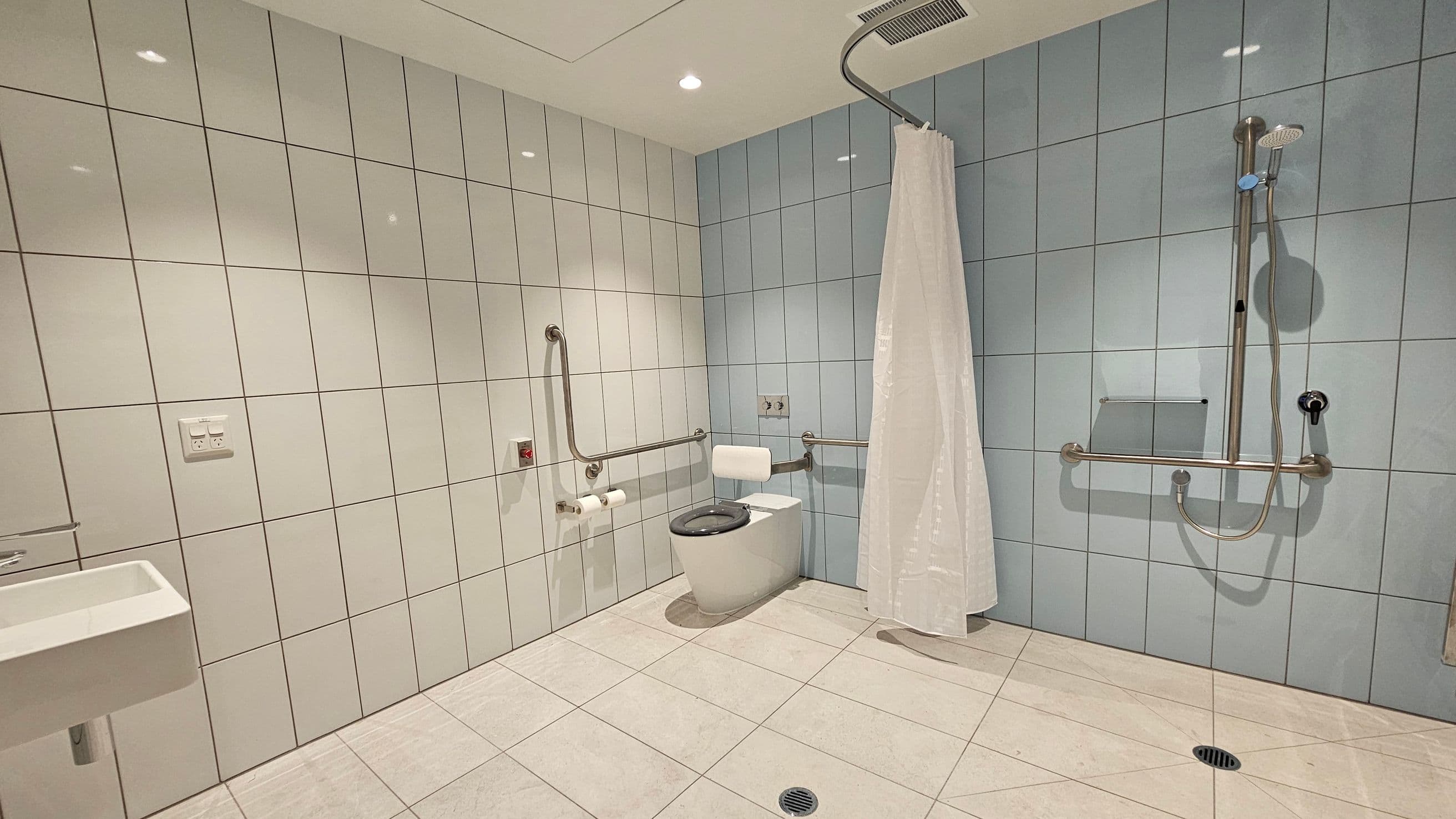 A spacious, accessible bathroom with white and light blue tiled walls. On the left, a small white sink is mounted on the wall. In the centre, a toilet with a black seat is equipped with grab bars and a toilet paper holder. On the right, a shower area with a curtain and a handheld showerhead is also fitted with grab bars. The floor is tiled in a light colour, and there are ceiling lights above.