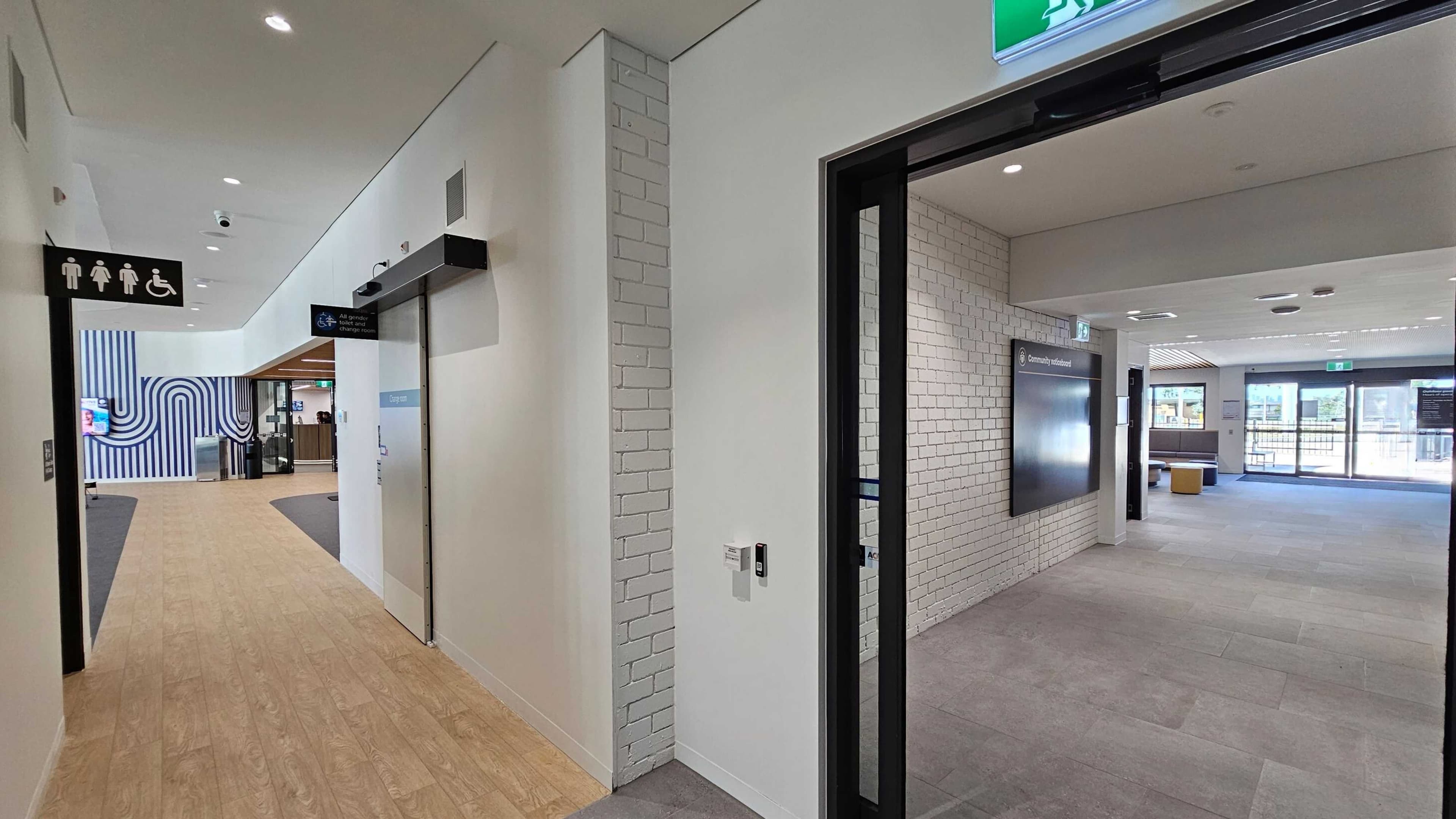 A modern indoor space with a corridor on the left featuring light wood flooring and a sign indicating toilets and accessibility. The wall has a blue and white striped design. On the right, there's a room with grey carpet tiles and white brick walls. A digital screen is mounted on the wall, and a person stands near large glass doors leading outside. The ceiling has recessed lighting and an exit sign above the door.