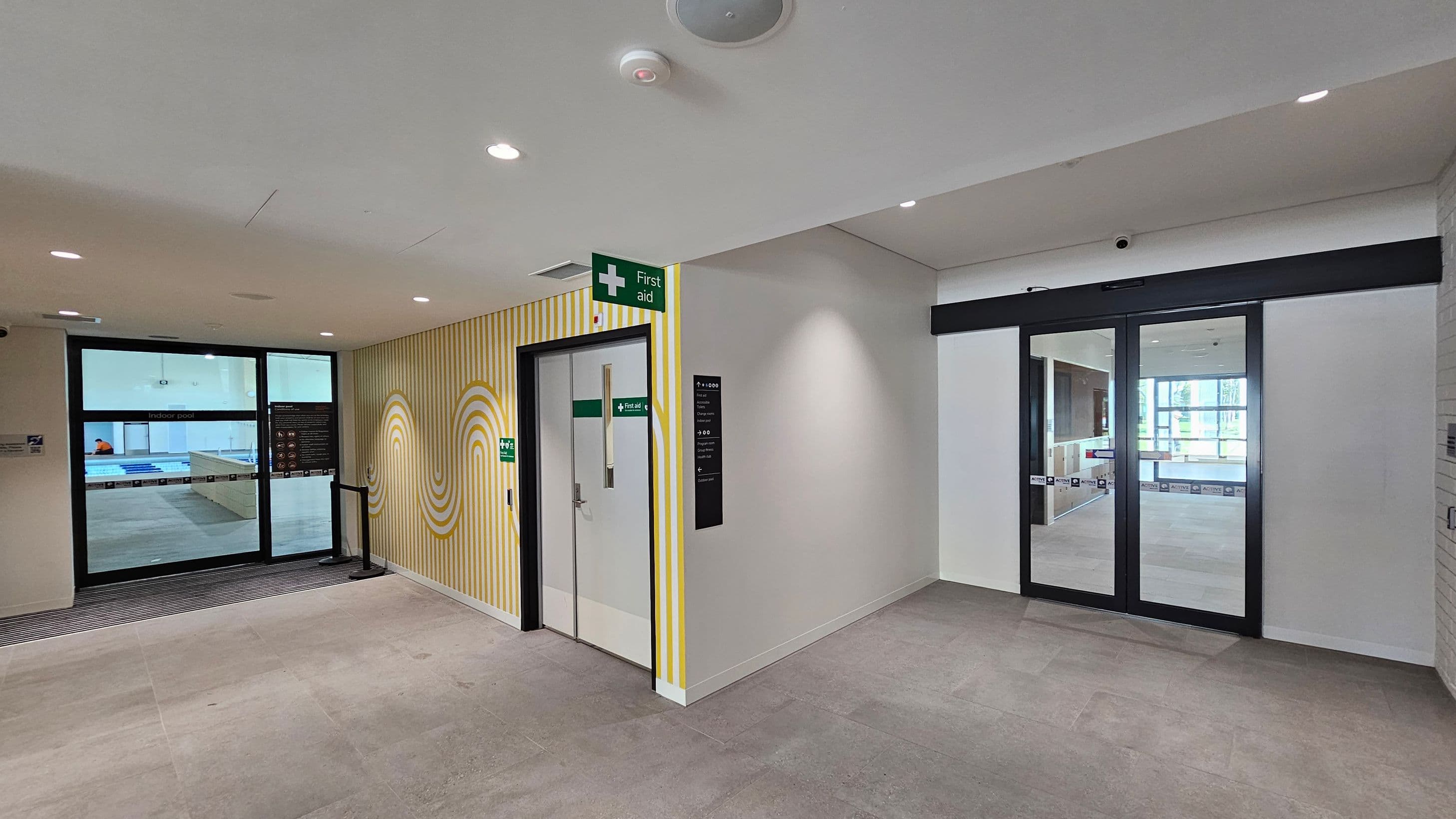 A hallway with light-coloured flooring and a white ceiling with recessed lights. On the left, there is a glass door leading to an indoor pool area, with a sign above it. In the centre, a door marked "First Aid" is set in a wall with yellow and white striped patterns. On the right, there are glass double doors with a black frame, leading to another room with large windows. A small black sign is mounted on the wall near the first aid door.