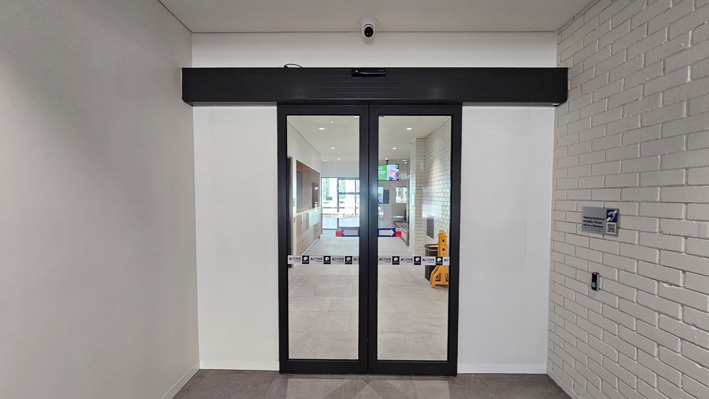 A pair of glass doors with a black frame is centred in the image, leading to a hallway with a tiled floor. Above the doors is a security camera. The left wall is plain white, while the right wall is made of white bricks and features a sign and a card reader. Through the doors, a corridor with a light-coloured floor and walls is visible, along with a digital screen and a yellow caution sign.