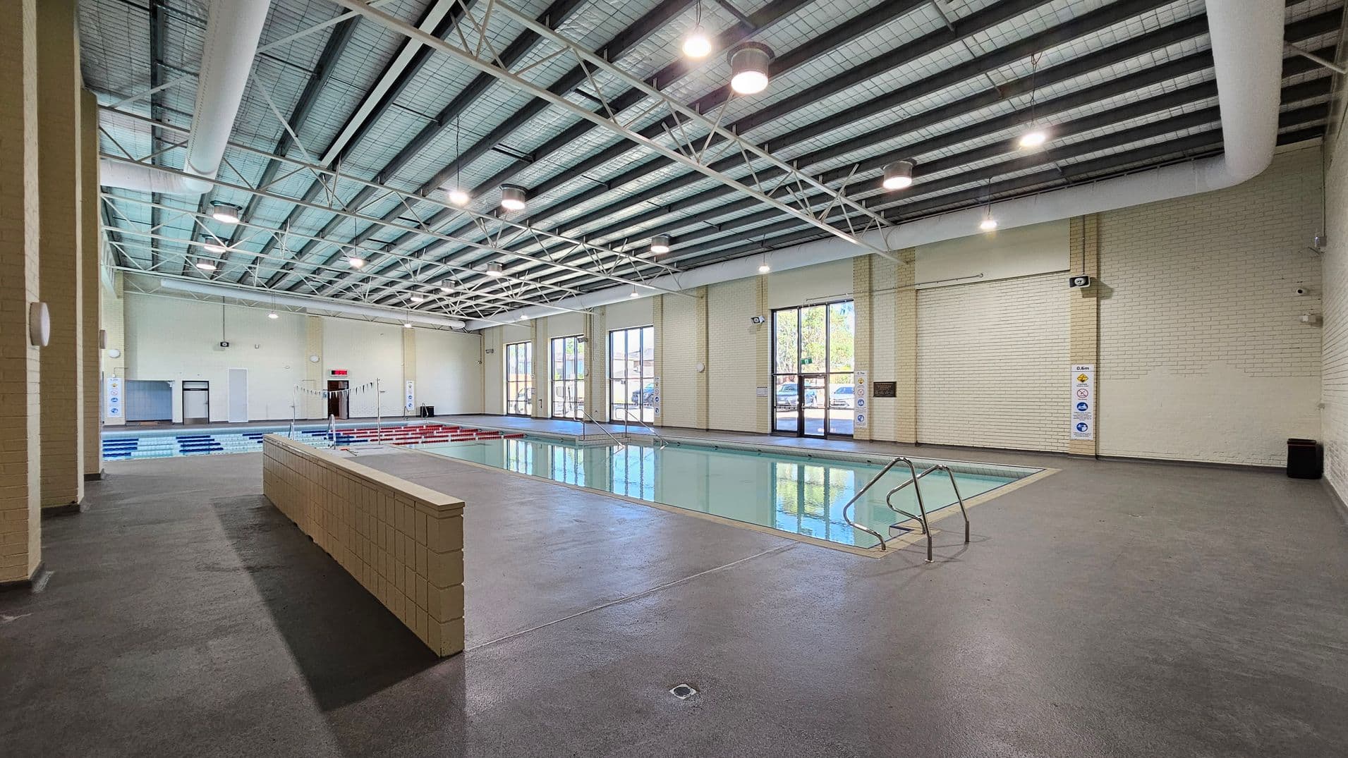 An indoor swimming pool with a light blue tiled floor and beige brick walls. On the left, there is a low brick wall separating the pool area from the walkway. The pool has metal handrails on the right side. Large windows line the right wall, allowing natural light to enter. The ceiling features exposed beams and hanging lights. The floor is a textured grey surface, and there are safety signs on the walls.