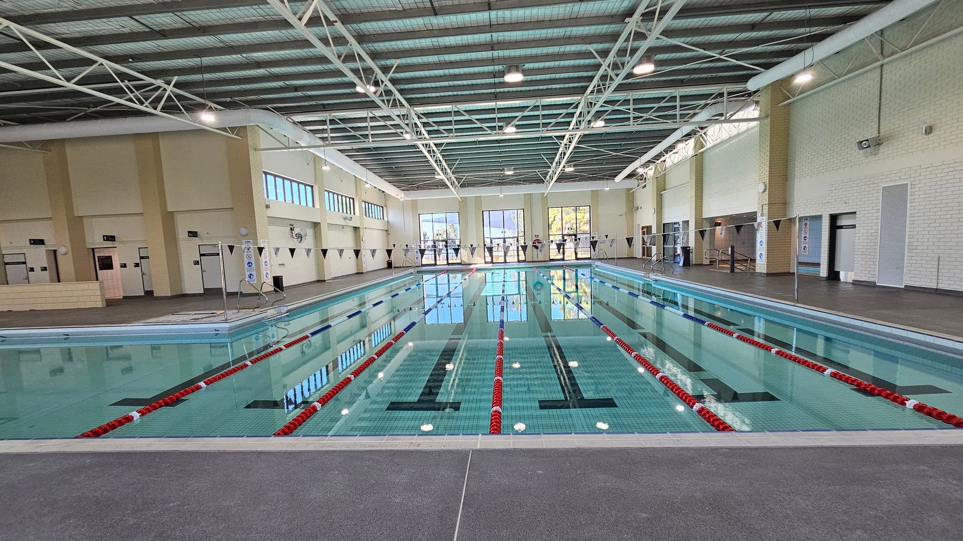 An indoor swimming pool with multiple lanes, separated by red and blue lane dividers. The pool is surrounded by a grey tiled floor. On the left, there are beige walls with several doors and a small counter. The right side features white brick walls with more doors. Large windows at the far end allow natural light to enter, and the ceiling is supported by metal beams with overhead lights.