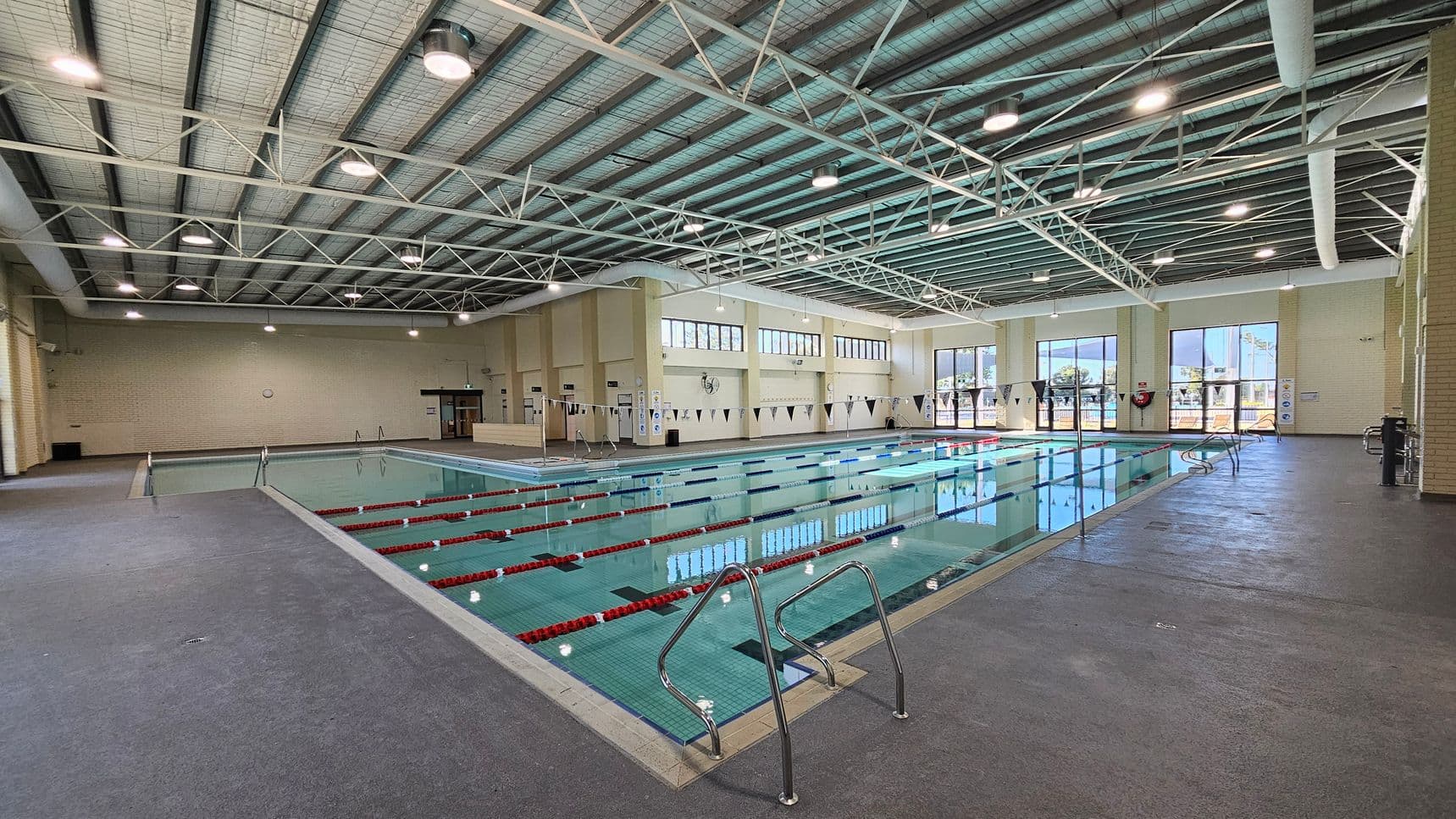 Indoor swimming pool with multiple lanes, marked by red lane dividers. On the left, a grey tiled floor surrounds the pool. The right side features large windows allowing natural light in, with views of trees outside. The ceiling is high with exposed beams and hanging lights. A metal ladder is at the pool's edge in the foreground. Walls are light-coloured with safety equipment visible.