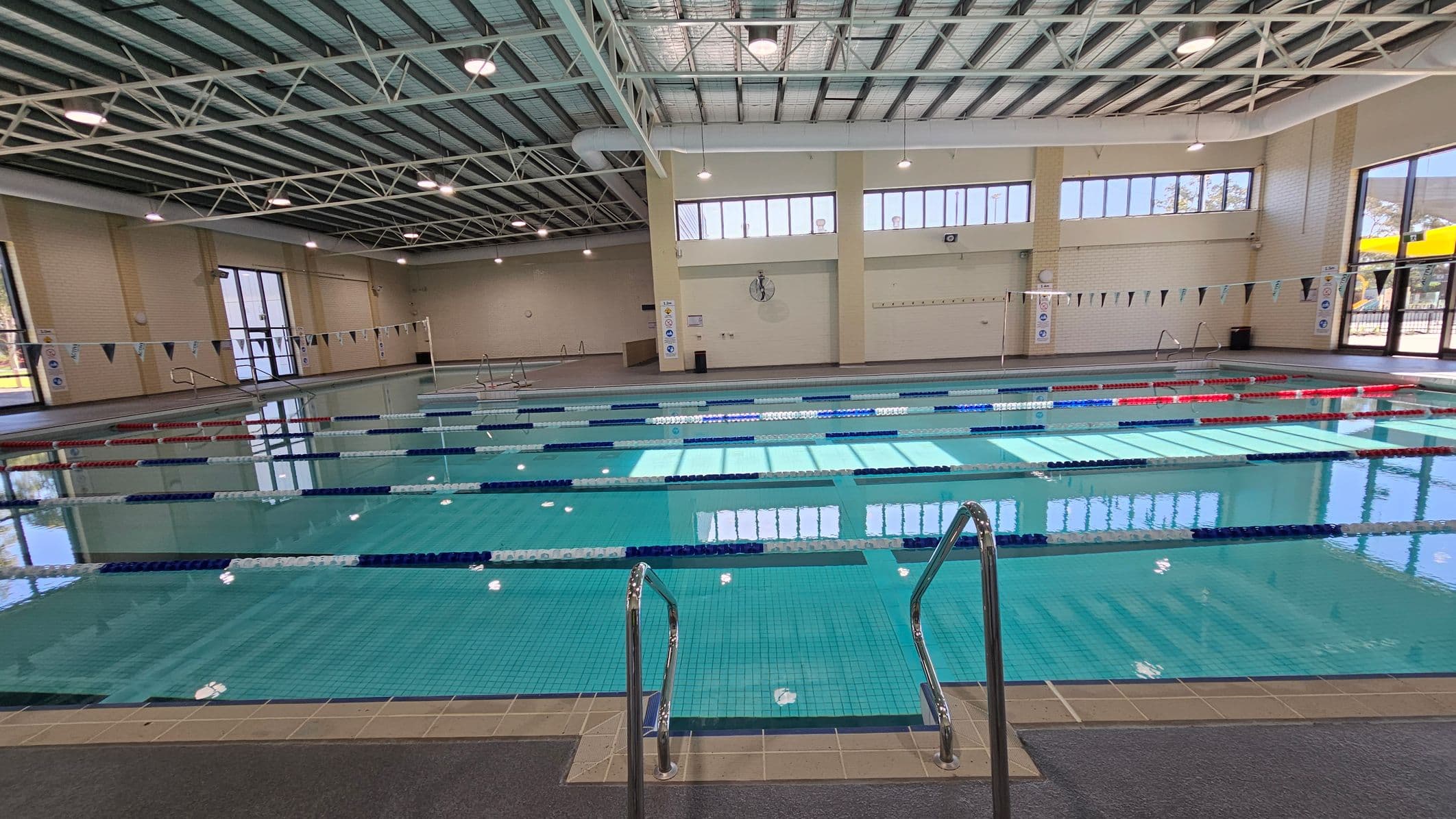 Indoor swimming pool with multiple lanes marked by blue, red, and white lane dividers. In the foreground, metal handrails lead into the pool. The background features large windows and beige walls with a clock and safety signs. The ceiling is high with exposed beams and lights. The floor around the pool is tiled.