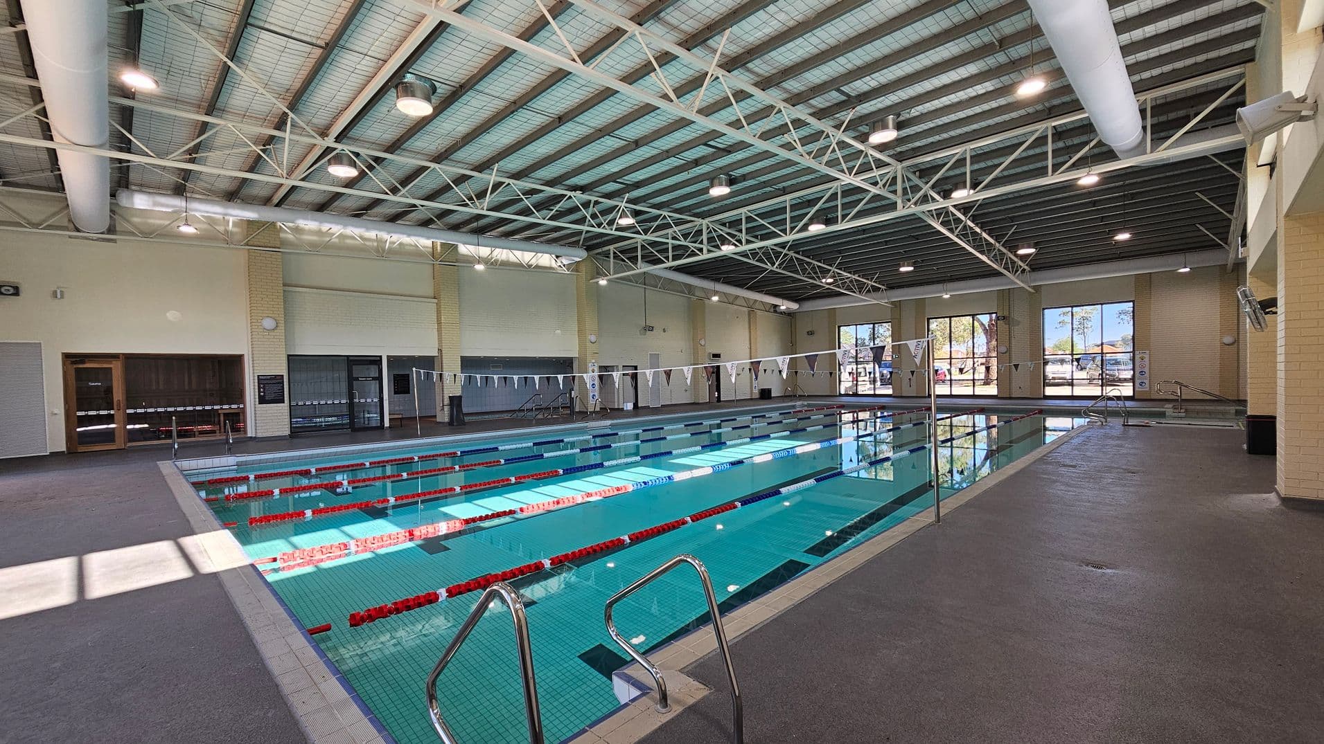 An indoor swimming pool with multiple lanes, marked by red and blue lane dividers. On the left, there are beige walls with large windows and a closed door. The right side features more windows, allowing natural light to illuminate the pool area. The ceiling is high with exposed beams and lighting fixtures. The floor surrounding the pool is grey and textured for safety. In the background, there are trees visible through the windows.