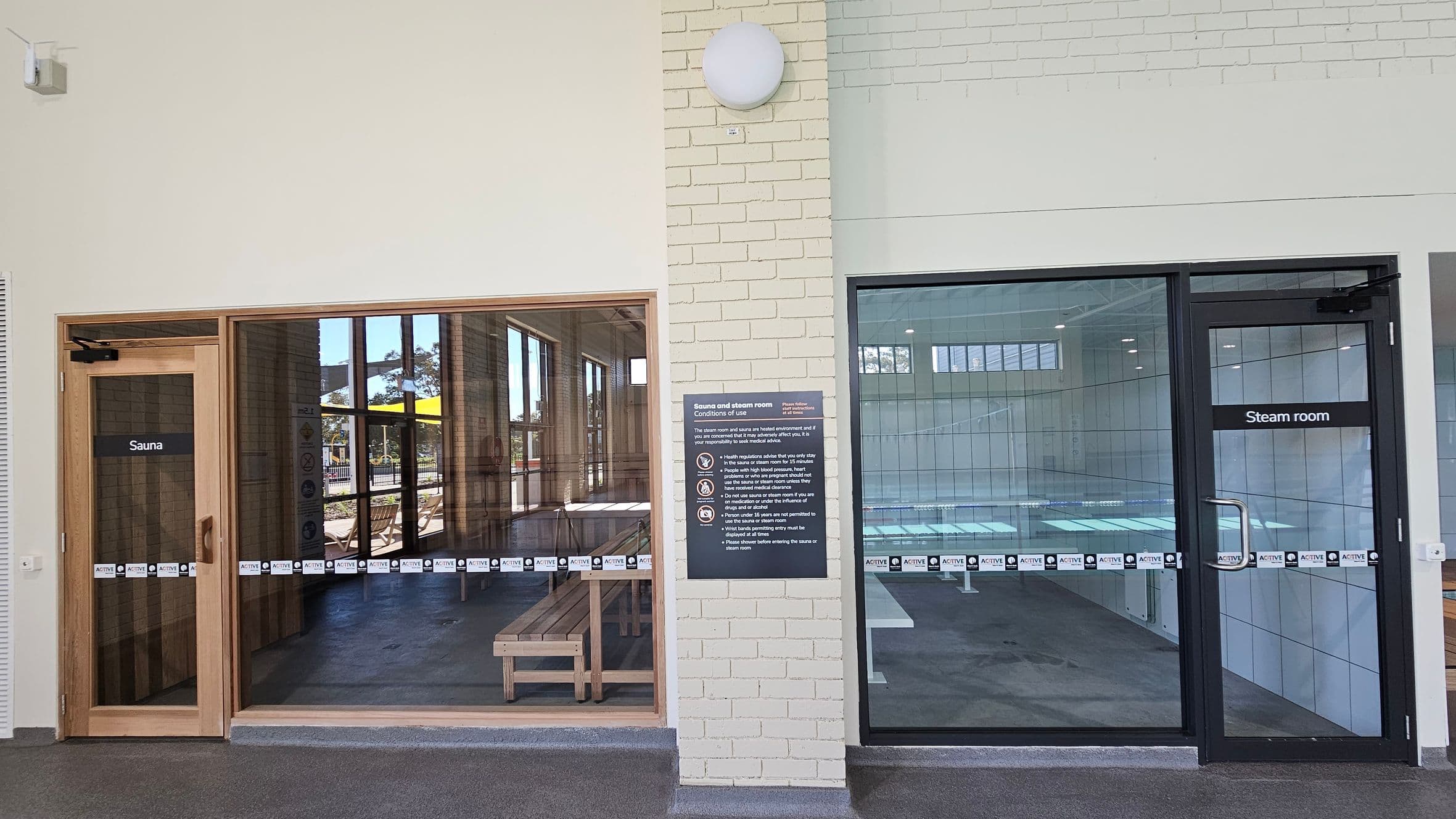 On the left, there is a wooden-framed glass door labelled "Sauna," with a view of wooden benches inside. On the right, a glass door labelled "Steam room" reveals a tiled interior. In the centre, a sign on a light brick wall outlines the conditions of use for the sauna and steam room. The floor is covered with dark grey carpet tiles, and the ceiling is not visible.
