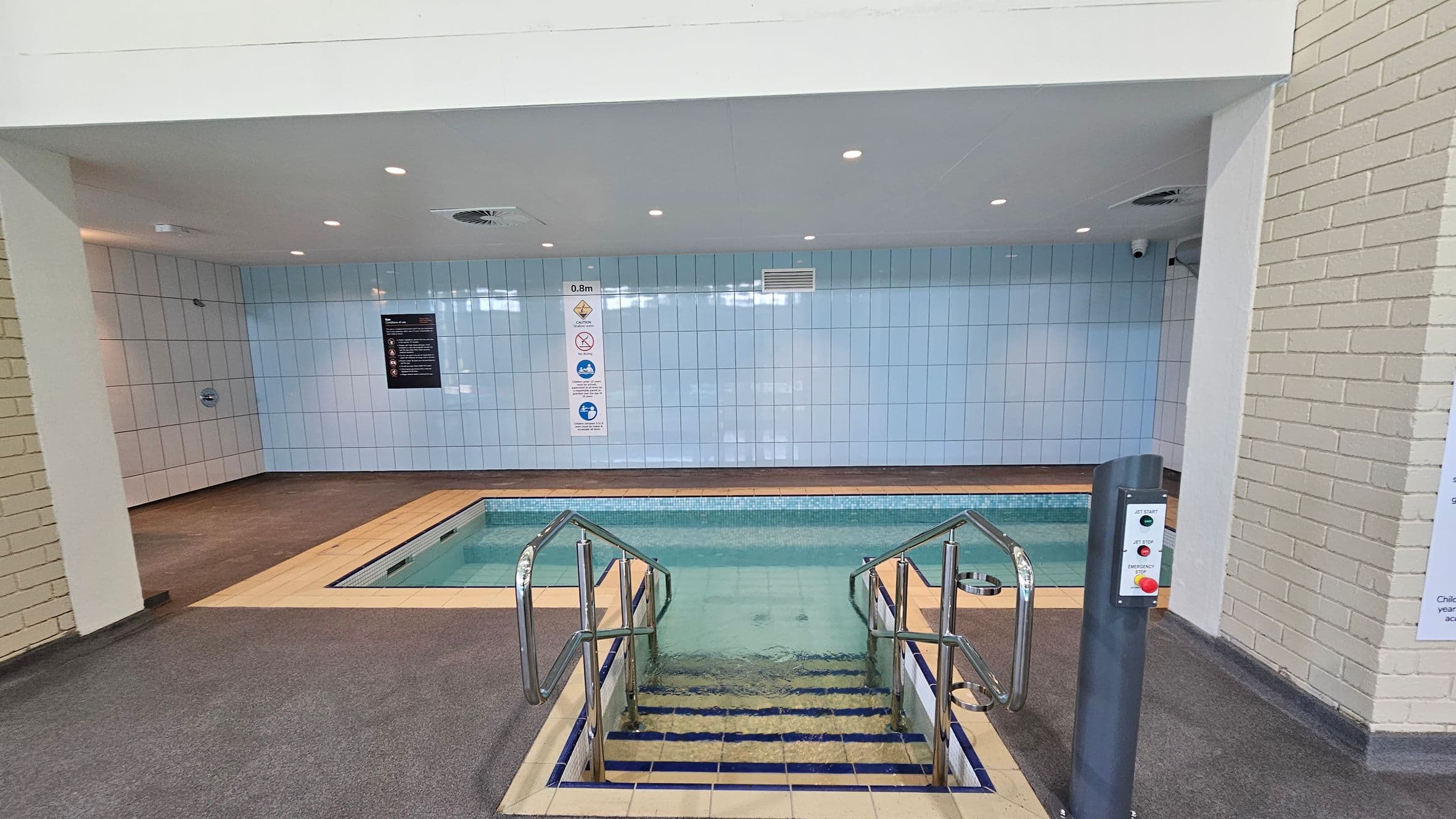 An indoor pool area with light blue tiled walls and a small rectangular pool. The pool has metal handrails and steps leading into the water. On the right, there's a control panel with buttons labelled "Jet Start" and "Jet Stop." The floor is covered with grey carpet, and the ceiling is white with recessed lighting. Signs with safety instructions are visible on the walls.