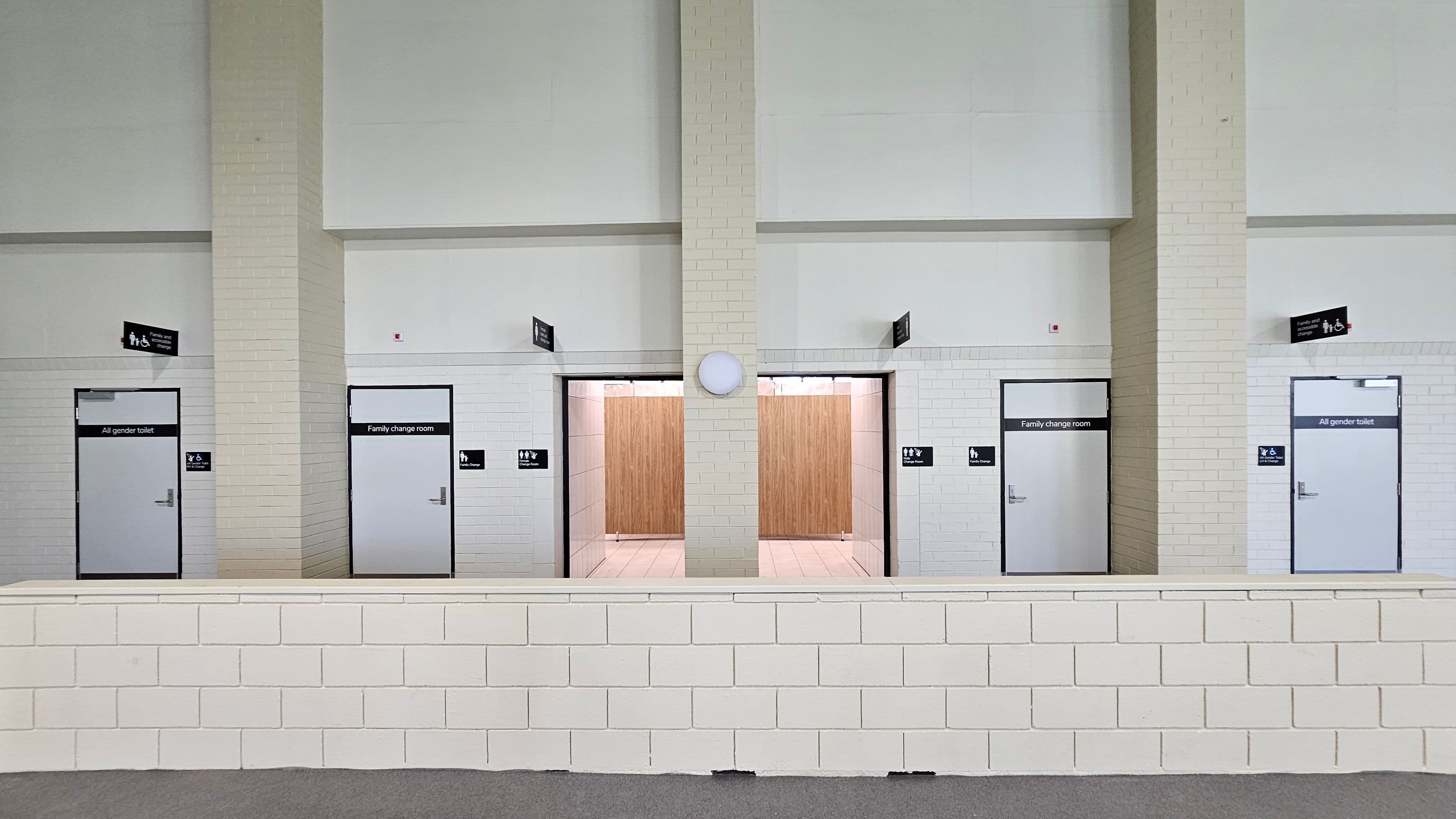 A row of doors set in a white brick wall. From left to right, there is a door labelled "All gender toilet," followed by an open entrance to a "Family change room" with wooden partitions visible inside. Next is a closed door also labelled "Family change room," and another door marked "All gender toilet." Above each door are black signs with icons. A low white brick wall runs horizontally in the foreground. The ceiling is high and plain.