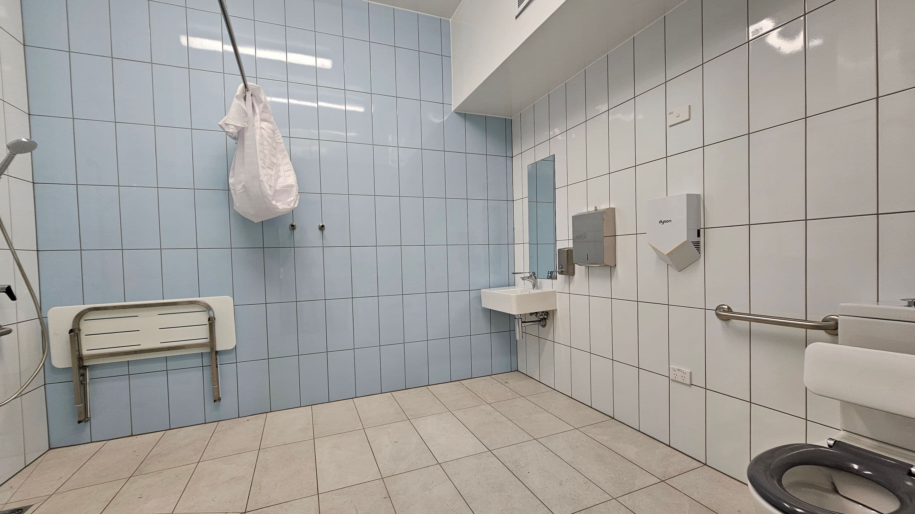 A spacious, accessible bathroom with light grey tiled walls and beige tiled floor. On the left, there's a foldable shower seat and a handheld showerhead. In the centre, a white sink with a mirror above it is mounted on the wall. To the right, a toilet with a black seat and a grab bar is visible. A plastic bag hangs from a hook on the wall, and a hand dryer and paper towel dispenser are mounted near the sink. The ceiling is white with fluorescent lighting.