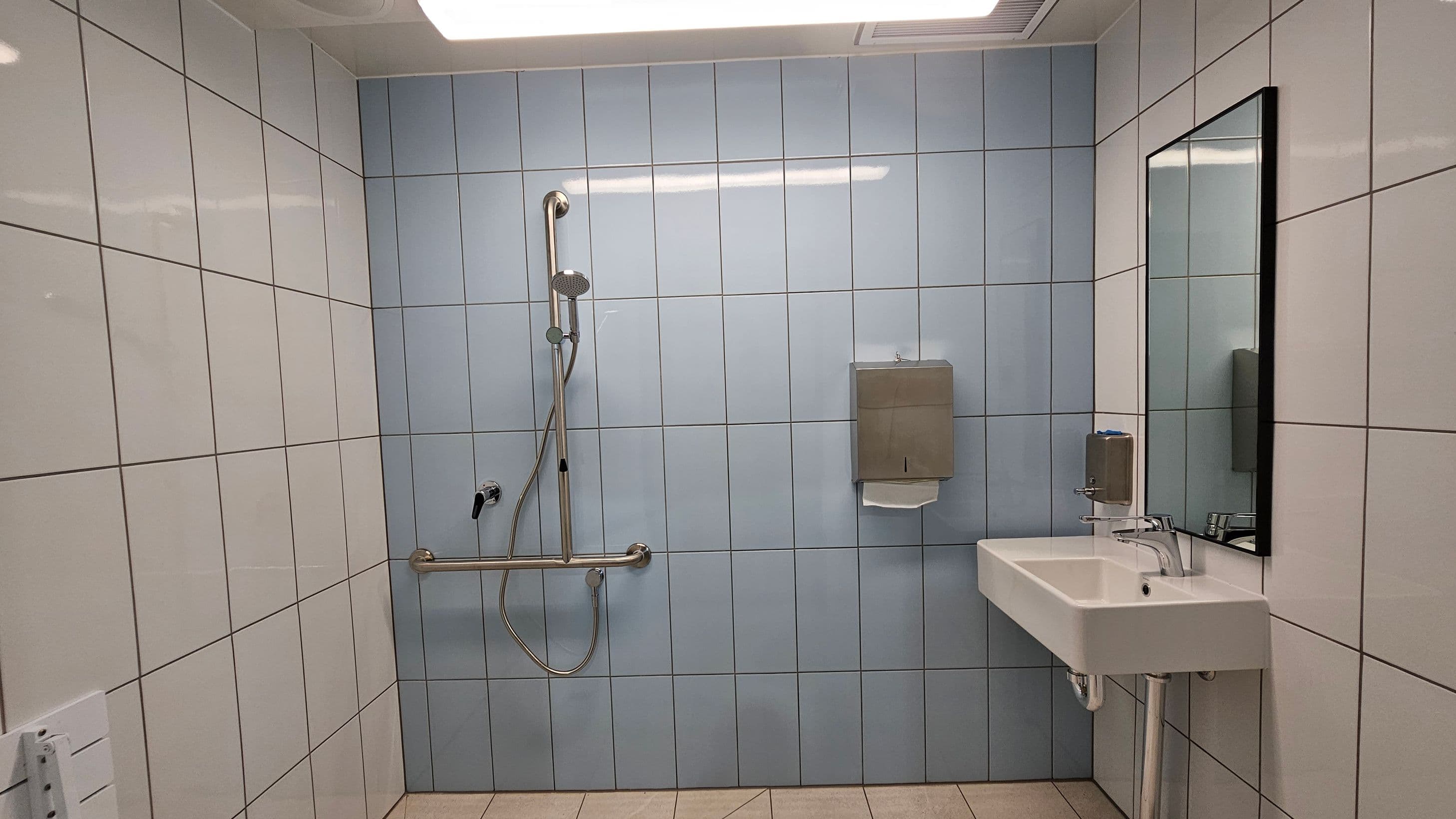 A bathroom with light grey tiled walls and a beige tiled floor. On the left, there is a shower area with a handheld showerhead and a horizontal grab bar. In the centre, a paper towel dispenser is mounted on the wall. On the right, a white sink with a chrome tap is below a rectangular mirror. A soap dispenser is attached to the wall next to the sink. The ceiling is white with a rectangular light fixture.