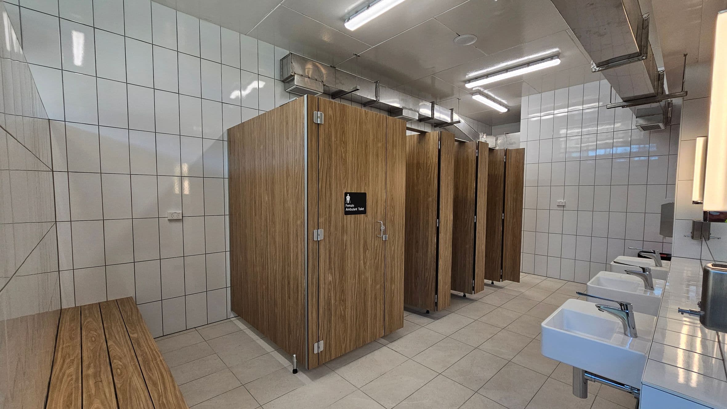 A public restroom with white tiled walls and beige tiled floor. On the left, there is a wooden bench. In the centre, there are several wooden cubicles with a sign indicating a female ambulant toilet on one door. On the right, a row of white sinks with chrome taps is mounted on a tiled counter. Above, fluorescent lights and exposed ductwork are visible on the ceiling.
