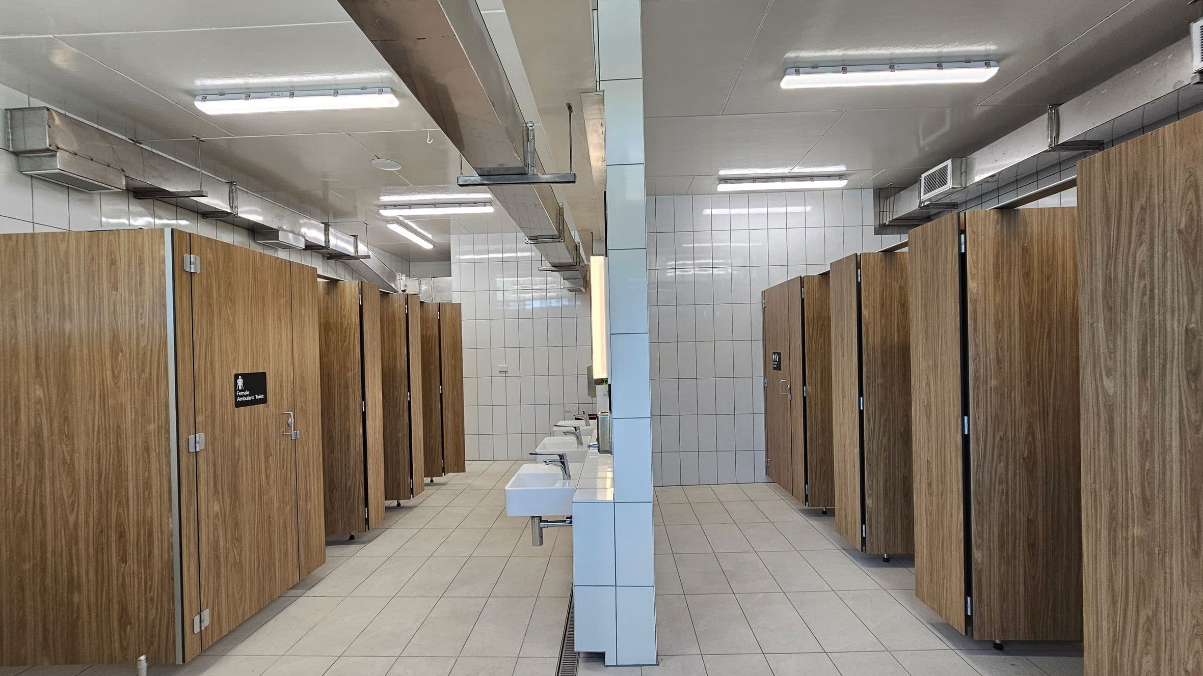 A public restroom with two rows of wooden cubicle doors on either side. The floor is tiled in light grey, and the walls are covered with white tiles. In the centre, there are white sinks mounted on a tiled partition. Overhead, fluorescent lights and metal ventilation ducts are visible on the white ceiling.