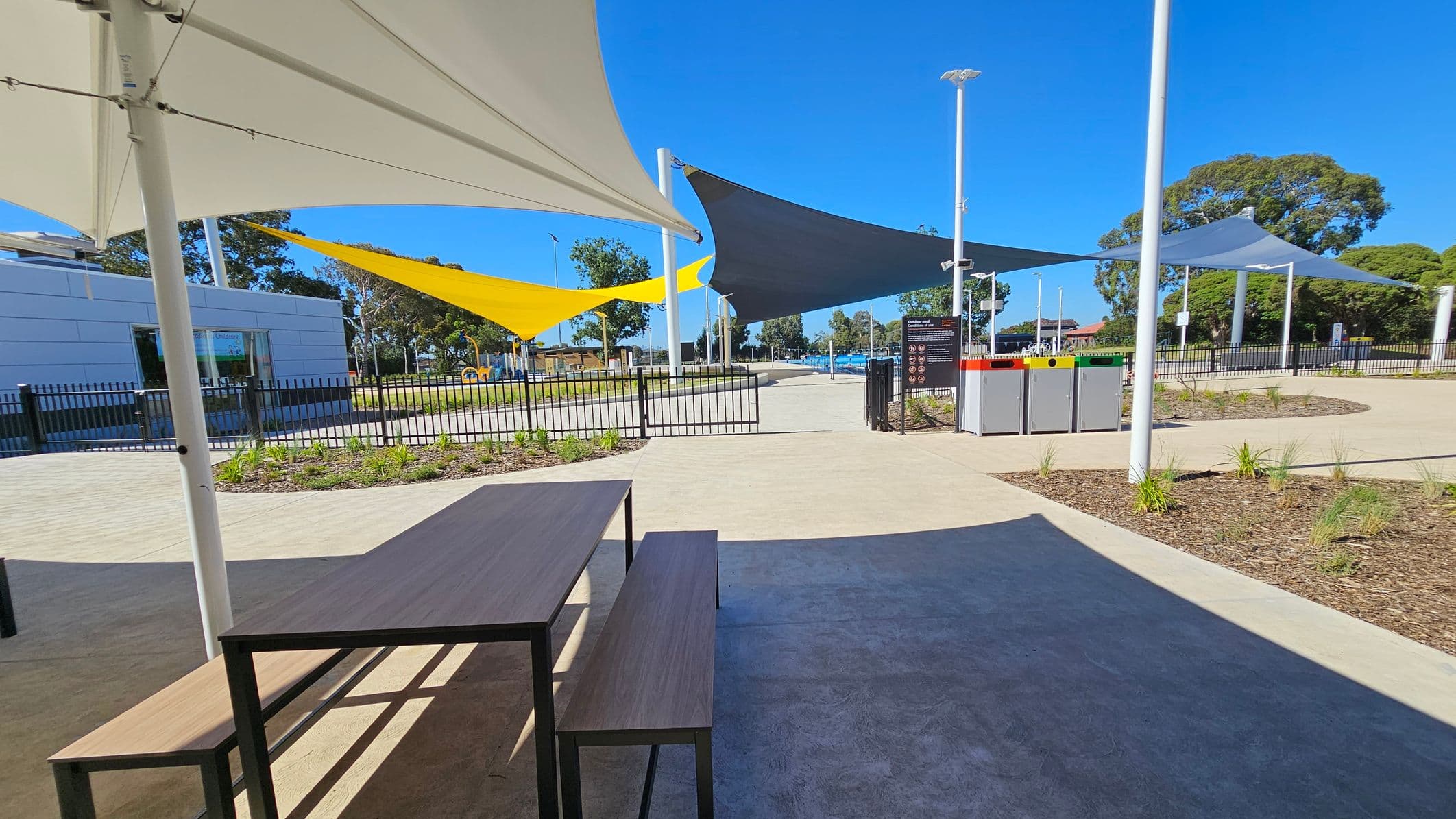 A park area with a wooden picnic table and benches on the left under a white shade sail. In the centre, there are yellow and dark grey shade sails. A black metal fence and a modern building with a large window are visible in the background. On the right, there are three waste bins with coloured lids next to a sign. The ground is paved with light and dark concrete, and there are small landscaped garden beds with young plants. Trees are scattered throughout the area.