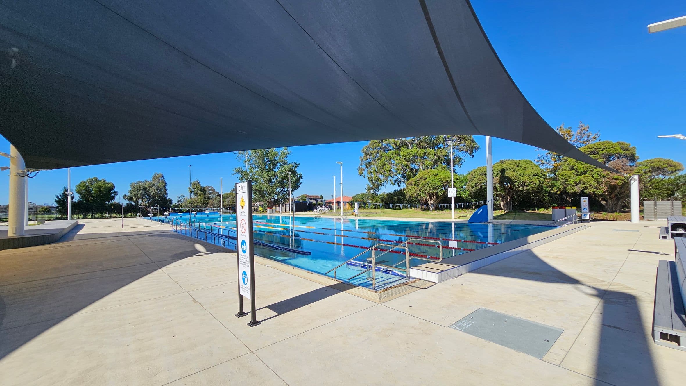 An outdoor swimming pool with clear blue water is in the centre, surrounded by a paved area. A large shade sail stretches across the top, casting shadows on the ground. On the left, there is a sign with pool rules. Trees and greenery are visible in the background, along with some seating areas. The pool has lane dividers, and there are steps leading into the water.