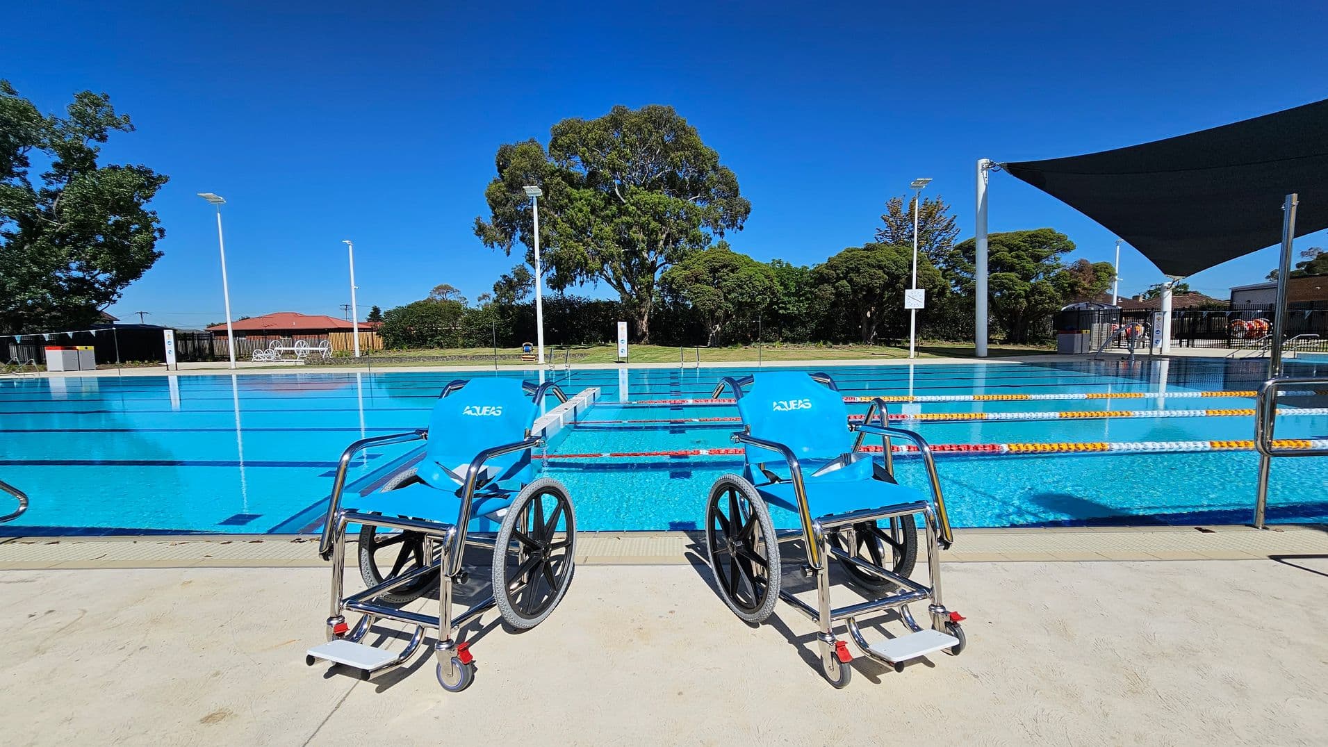 Two blue aquatic wheelchairs are positioned on a concrete poolside area. Behind them is a swimming pool with lane dividers in red and white. In the background, there are trees, a red-roofed building, and white outdoor furniture. A black shade sail is visible on the right, and several tall light poles are scattered around the area.