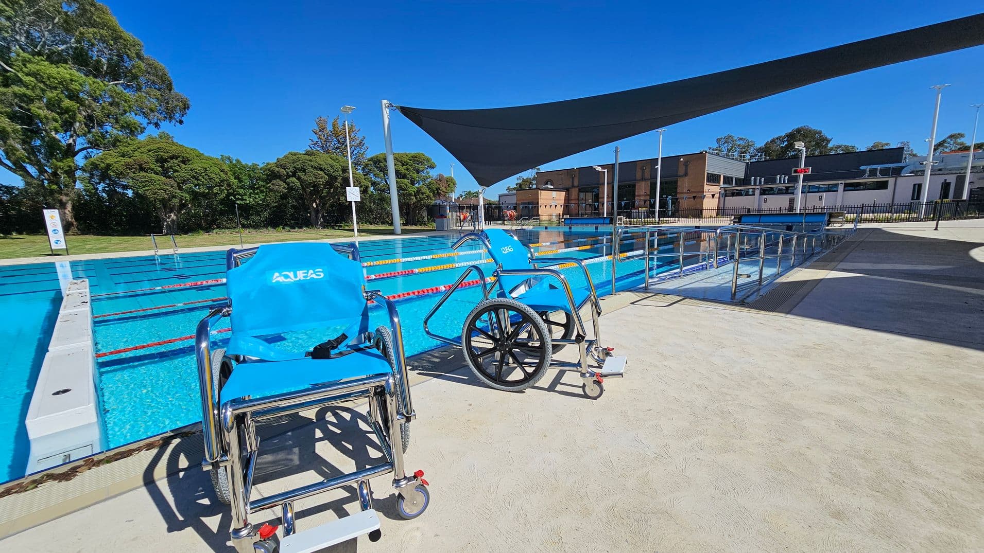 Two blue aquatic wheelchairs are positioned on a concrete poolside area. The pool has clear blue water with lane dividers. A shade sail is stretched above part of the pool. In the background, there are trees and a building with a flat roof. A metal railing runs along the pool's edge, and the ground is light-coloured concrete.