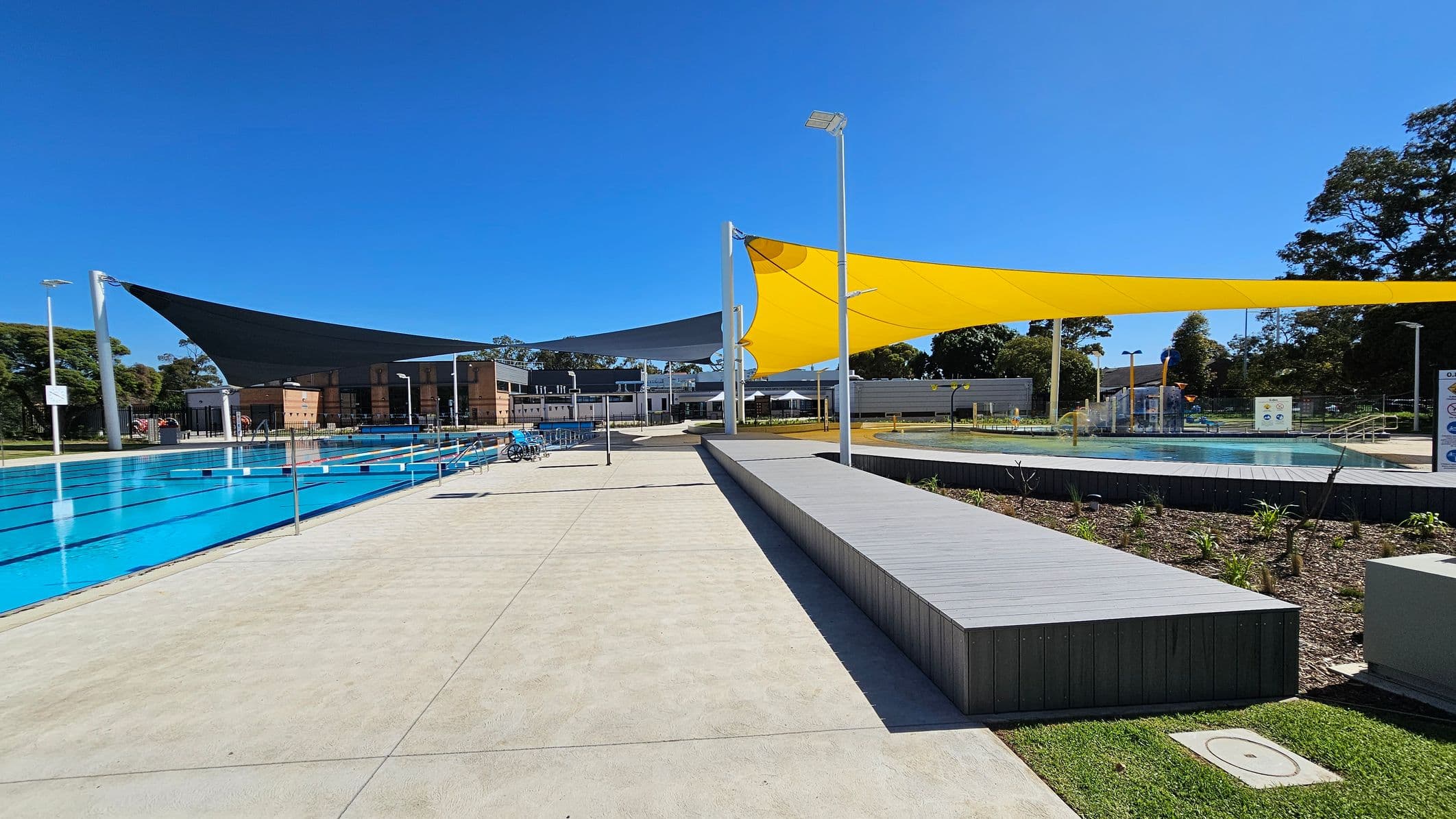 Outdoor swimming pool area with a large, rectangular pool on the left, featuring lane dividers. A modern building with wooden accents is in the background. Overhead, there are large triangular shade sails in black and yellow. On the right, a smaller pool with water features is visible, surrounded by landscaped gardens. The ground is paved with light-coloured concrete, and there are several tall light poles around the area. Trees are visible in the background.