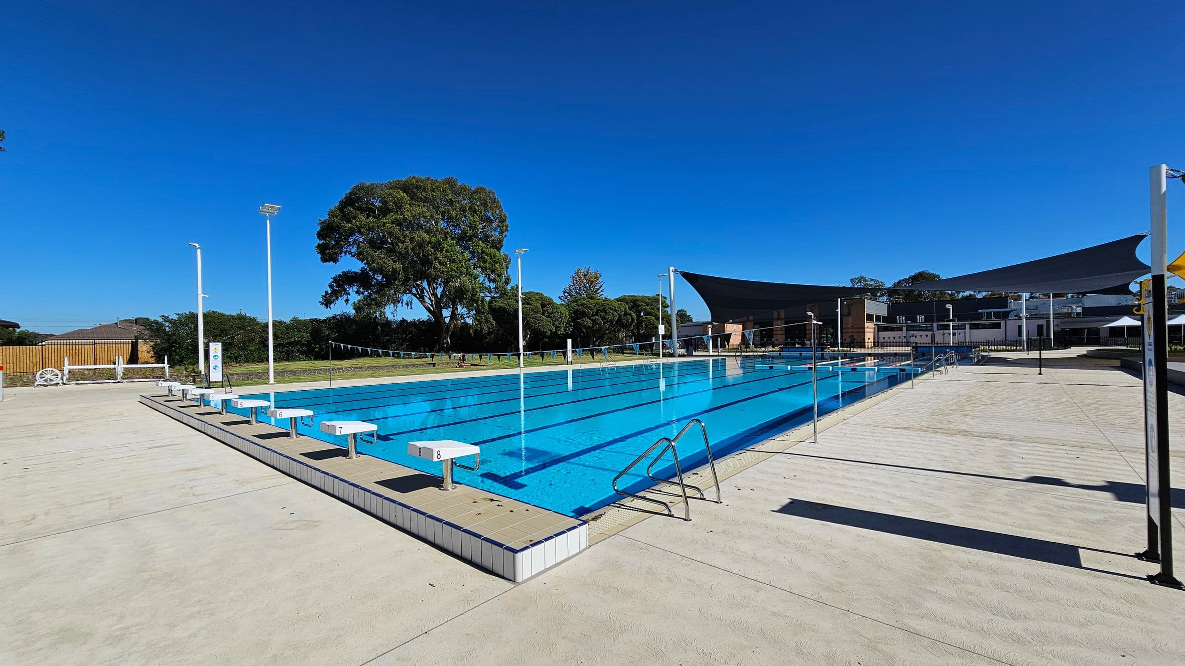 An outdoor swimming pool with clear blue water and marked lanes. Starting blocks are positioned at one end. The pool is surrounded by a light-coloured concrete deck. On the right, there are shade sails and a building in the background. Trees and bushes line the perimeter on the left, with tall light poles scattered around. A metal ladder is visible on the pool's edge.