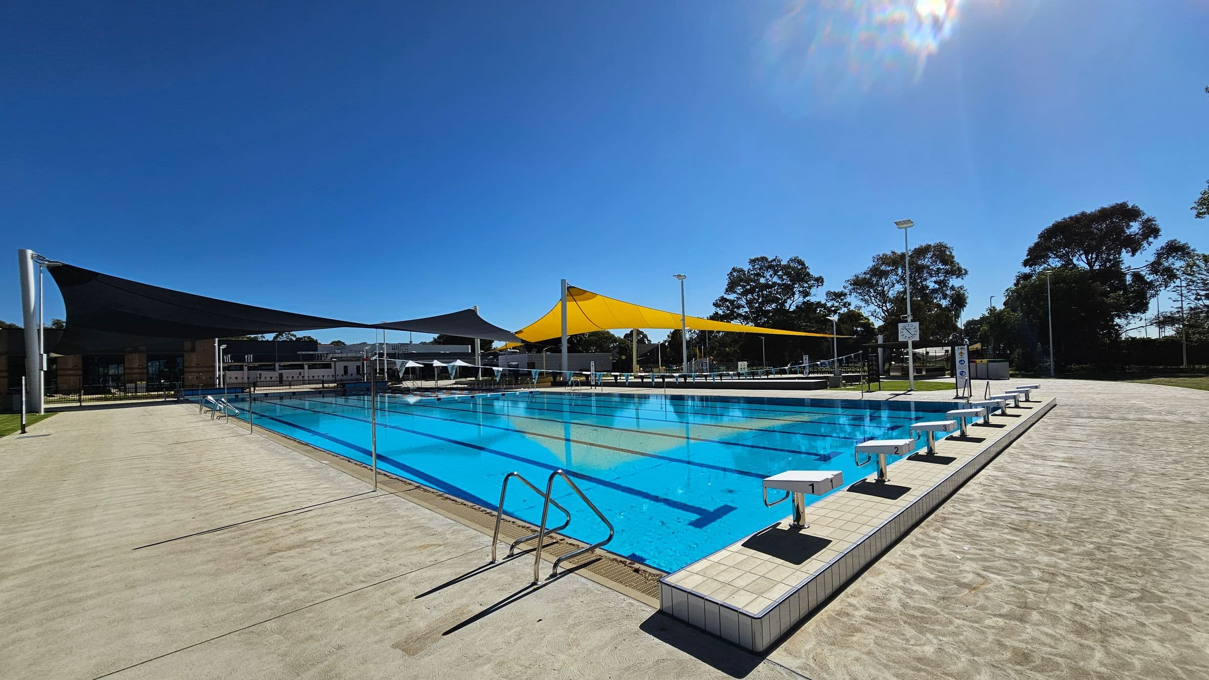 An outdoor swimming pool with clear blue water is surrounded by a paved area. On the left, there are large triangular shade sails in black and yellow, supported by metal poles. On the right, a row of starting blocks lines the pool's edge. In the background, trees and a few buildings are visible. The sky is clear and sunny.