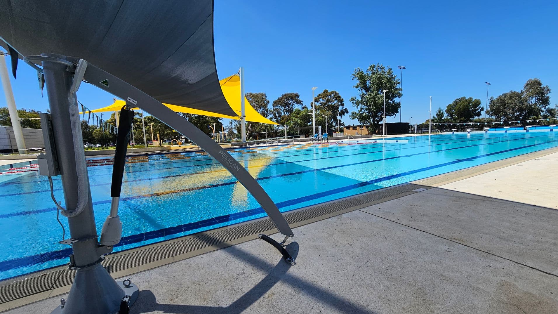 An outdoor swimming pool with clear blue water and lane dividers. On the left, a large grey structure supports a yellow shade sail. The pool deck is concrete, and there are trees and a few buildings in the background. The sky is clear and blue.