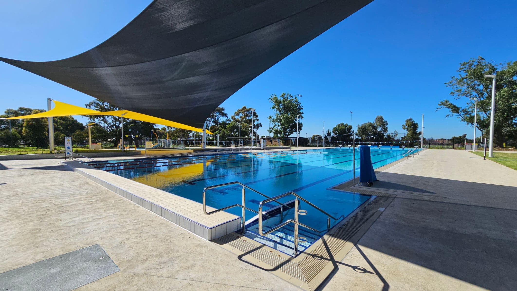 An outdoor swimming pool with clear blue water is in the centre, surrounded by a paved area. On the left, there are steps leading into the pool. Above, large black and yellow shade sails provide cover. In the background, there are trees and a fence enclosing the area. The ground is a mix of light and dark paving, and there are several tall light poles around the pool.