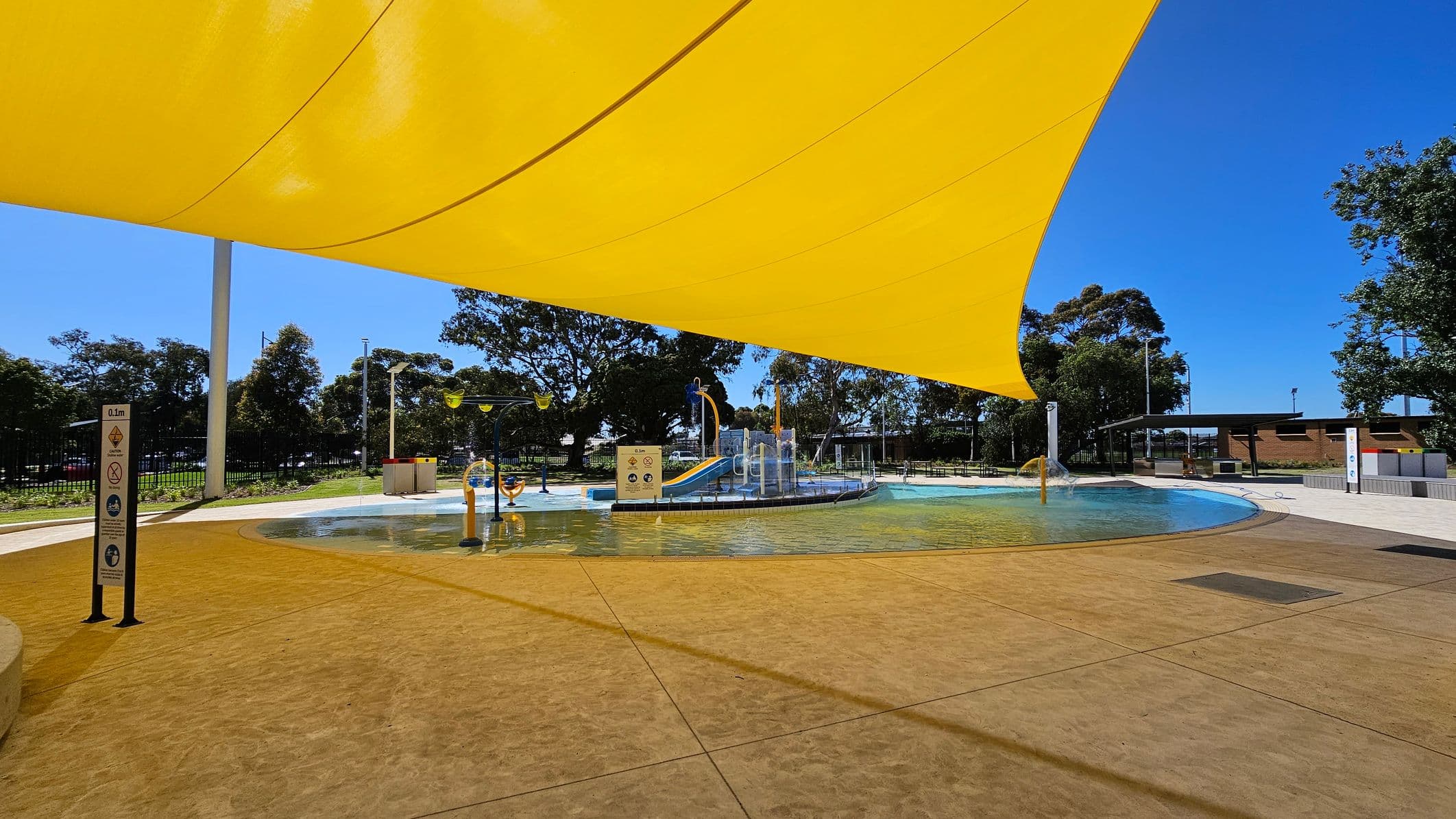 A children's splash park with a shallow pool and play equipment, including a slide and water features, is covered by a large yellow shade sail. The ground is paved with a textured surface. Trees and a fence line the background, with a small building and seating area to the right. The sky is clear and blue.