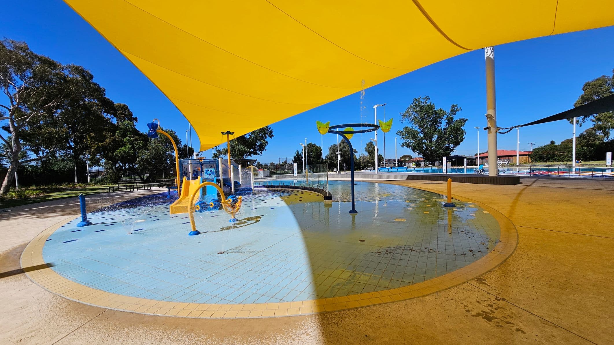 A children's splash pad with water play equipment, including a yellow and blue structure and a circular water feature with buckets. The ground is covered with light blue and yellow tiles. A large yellow shade sail covers part of the area. Trees and a fence are visible in the background, with a pool area to the right. The floor is a mix of wet and dry surfaces.