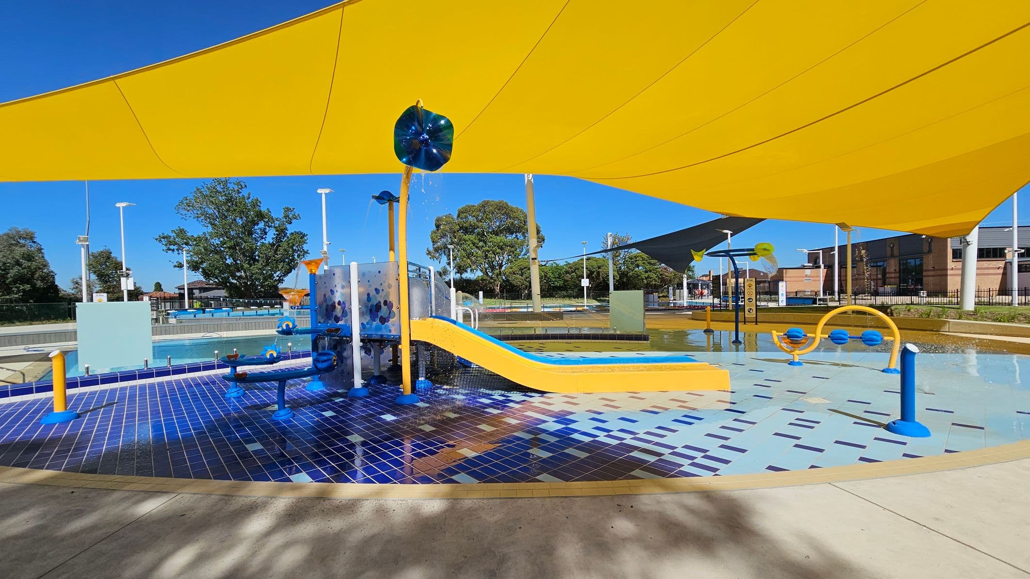A children's water play area with a blue and yellow theme. In the centre, there's a yellow slide leading into a shallow pool. The ground is covered with blue and light blue tiles. Yellow shade sails are overhead, providing cover. Various water play structures, including fountains and sprayers, are scattered around. Trees and buildings are visible in the background, and the area is surrounded by a fence.
