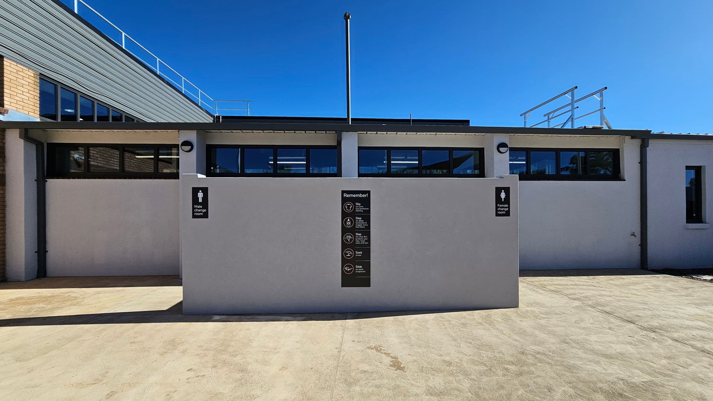 A building exterior with a light grey wall in the foreground, featuring signs for male and female change rooms on either side. In the centre, a black sign with safety reminders is visible. The building has a flat roof with a metal railing and several rectangular windows. The ground is a smooth concrete surface, and the sky is clear and blue.