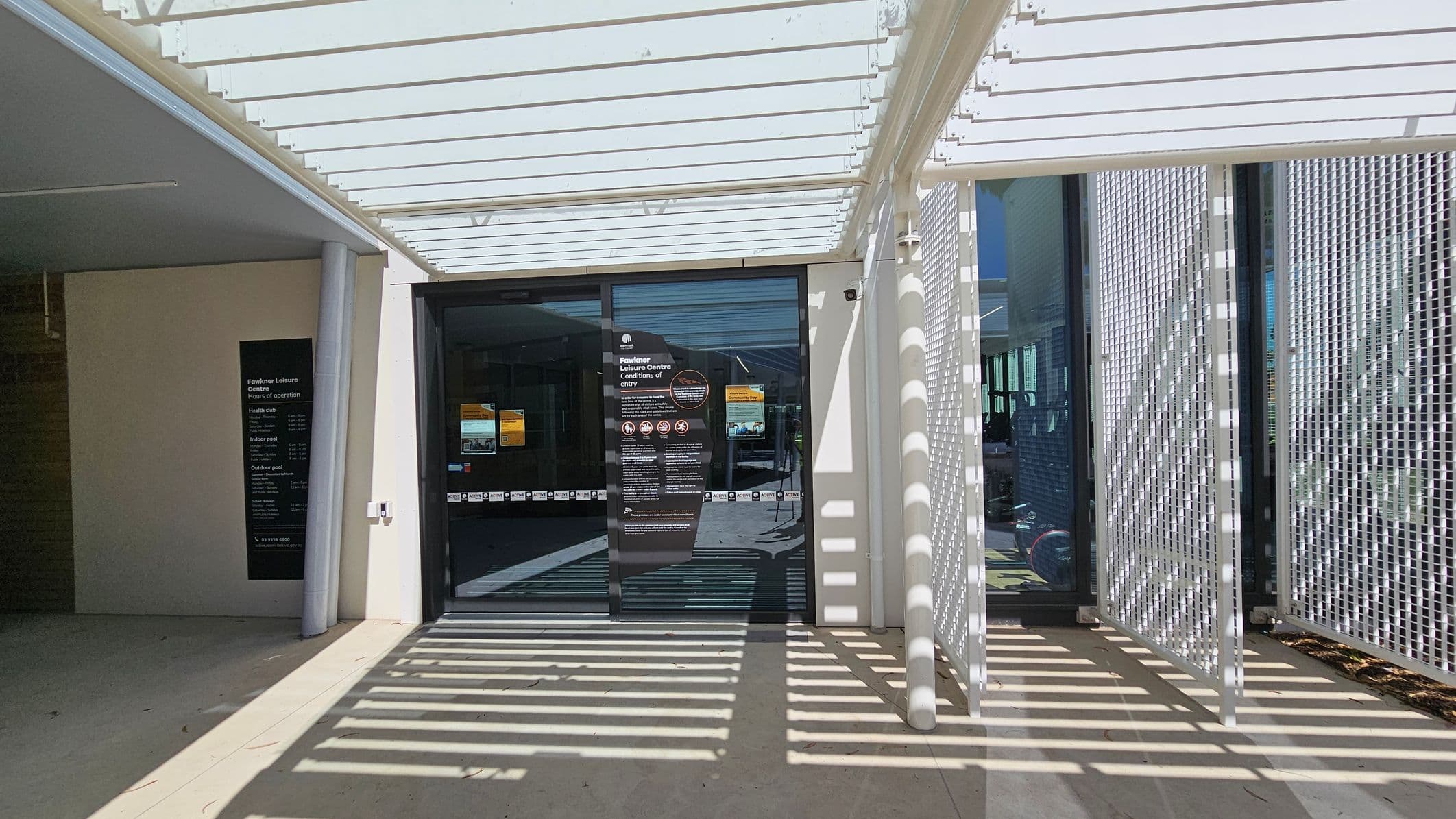 A glass entrance door with signage is in the centre, leading into a building. To the left, a wall displays a sign with hours of operation. The right side features a white lattice structure casting shadows on the ground. The ceiling consists of white slats, also casting shadows. The floor is light-coloured and smooth.