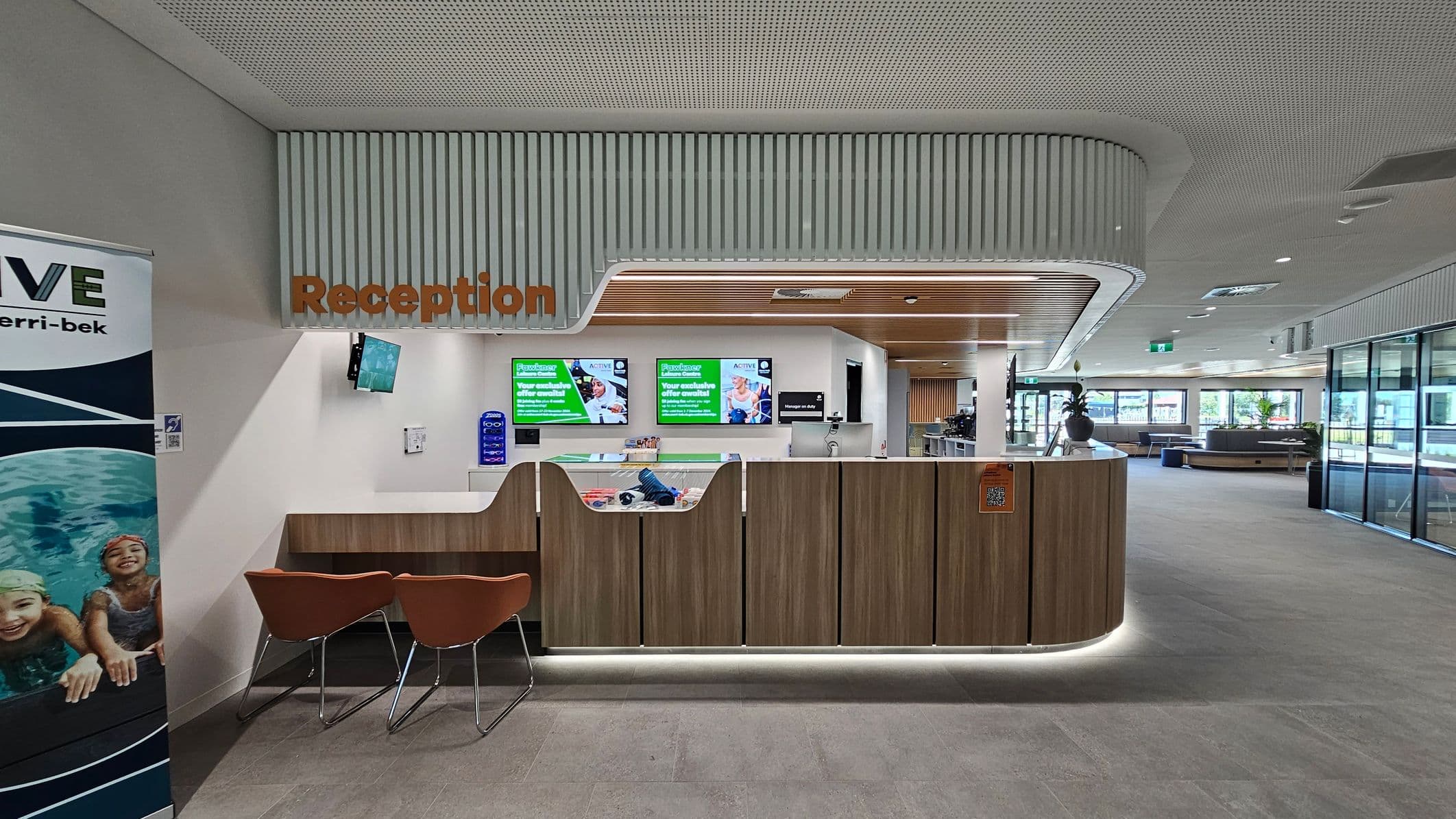 Reception area with a modern design. On the left, a banner with swimmers. The reception desk is centrally located, made of wood with a white countertop, and has two brown chairs in front. Above, the word "Reception" is displayed in orange. Behind the desk, digital screens show promotional content. The ceiling has a perforated design with wooden accents, and the floor is grey tile. To the right, large windows and seating areas are visible.