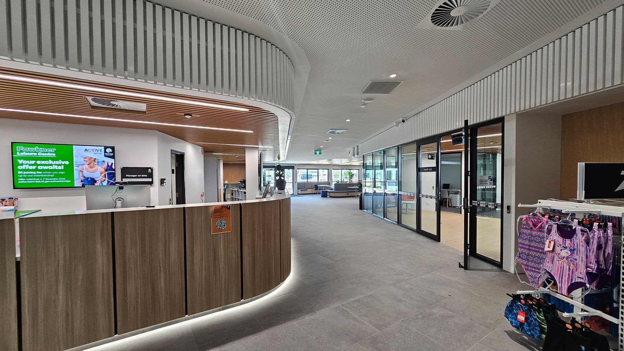 A modern interior with a curved wooden reception desk on the left, featuring a digital screen displaying information. The ceiling has a perforated design with integrated lighting. On the right, there are glass-walled rooms and a display rack with colourful swimwear. The floor is tiled in grey, and the space is well-lit with natural light coming from large windows in the background.