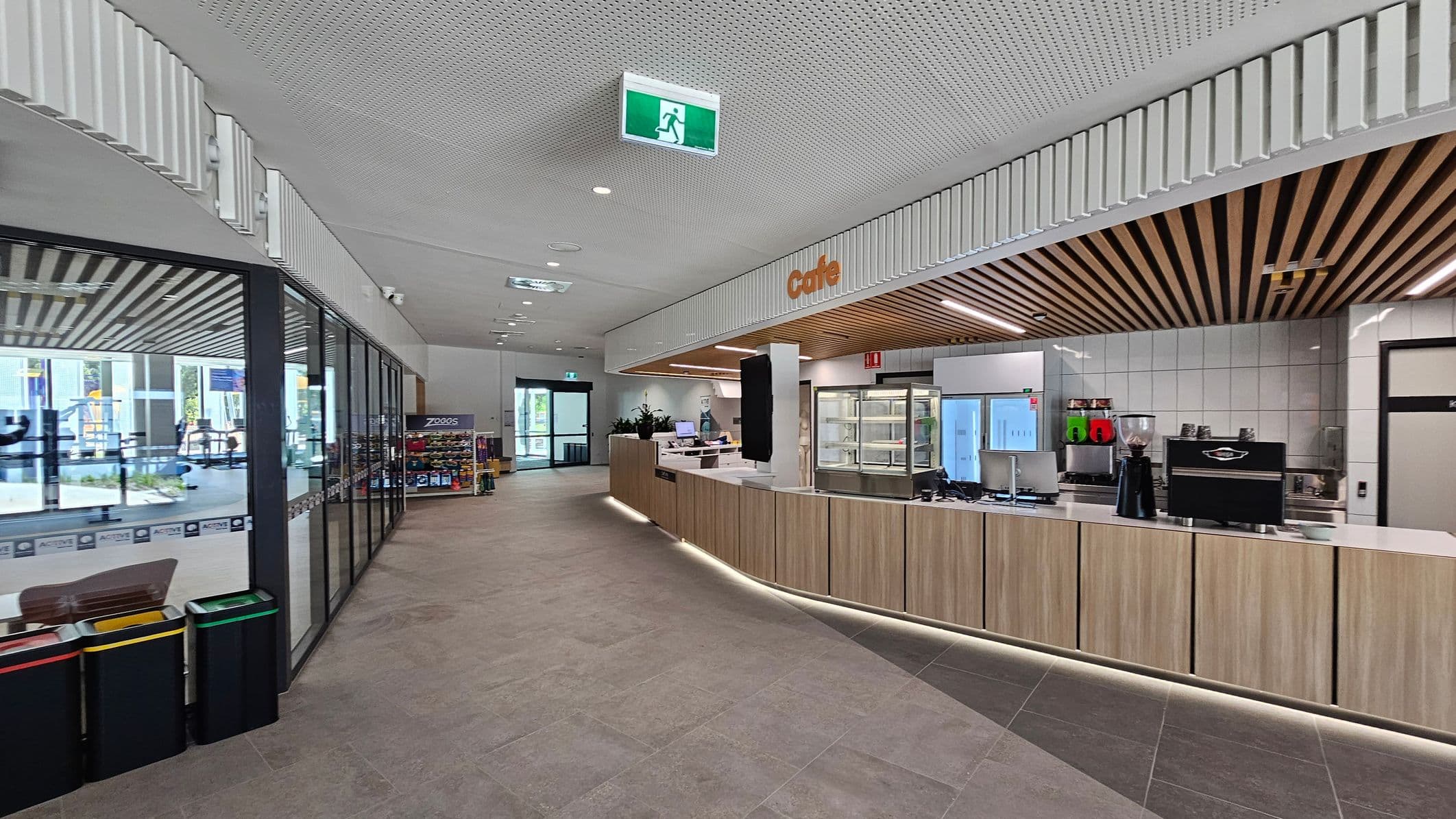 A modern indoor space with a café on the right, featuring a wooden counter and a coffee machine. Behind the counter are display fridges and shelves. The ceiling has a perforated design with wooden slats above the café area. On the left, there are large glass windows and doors, with a view of an adjacent area. Near the windows, there are recycling bins. The floor is tiled, and there are emergency exit signs on the ceiling.
