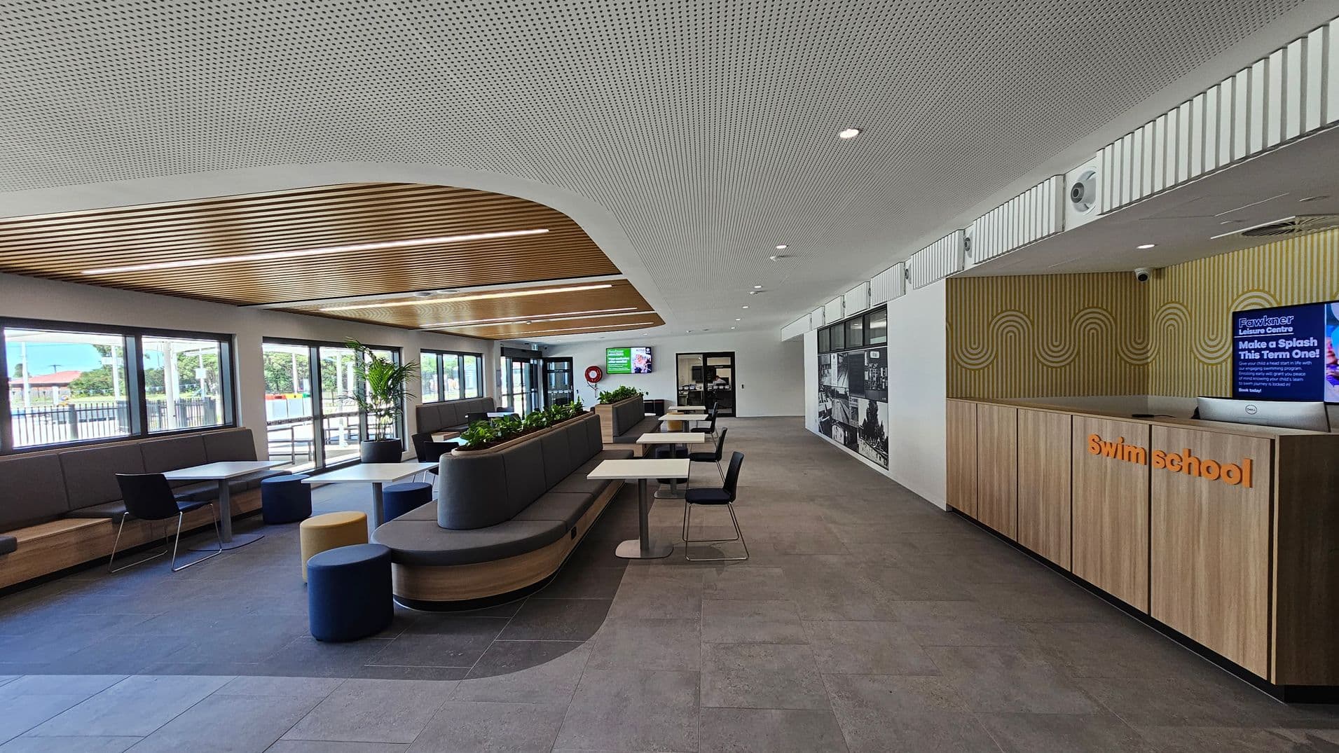 Modern interior of a leisure centre with a swim school reception on the right, featuring wooden panelling and a digital screen. In the middle, there are tables with chairs and a long, curved bench with planters. On the left, large windows allow natural light in, with more seating along the wall. The ceiling has a mix of perforated and wooden slats, and the floor is tiled in grey.