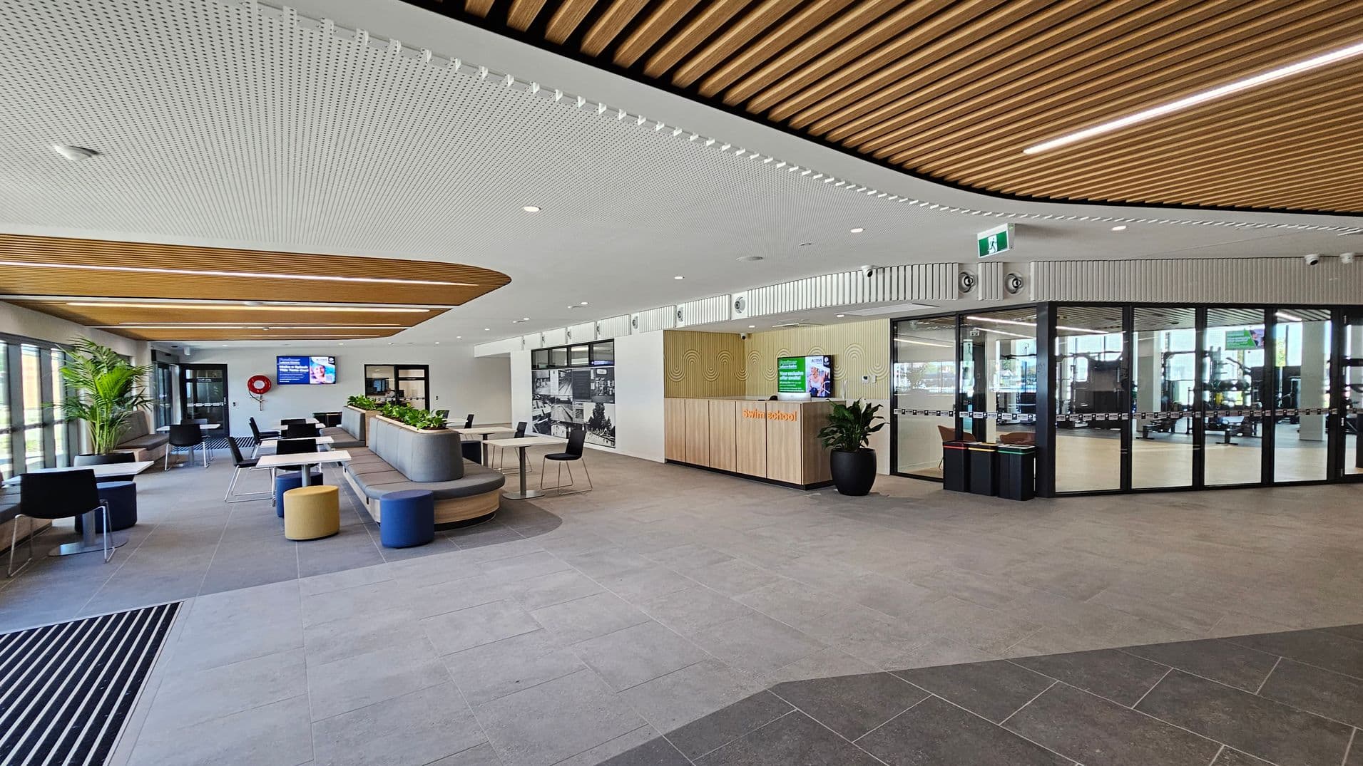 A modern interior space with a grey tiled floor and a ceiling featuring wooden slats and white perforated panels. On the left, there are large windows, a potted plant, and seating areas with tables and chairs. In the centre, a curved bench with planters is surrounded by stools. On the right, a reception area with a wooden counter and digital screen is adjacent to glass doors leading to another room.