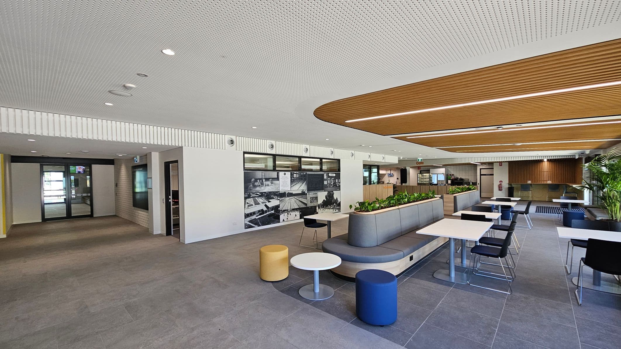 A modern interior space with a grey tiled floor and a perforated white ceiling. On the left, there are glass doors and a wall with black-and-white images. In the centre, there is a seating area with a curved bench, small round tables, and colourful stools. On the right, there are more tables with black chairs and a wooden ceiling feature. The background includes a counter area and potted plants.