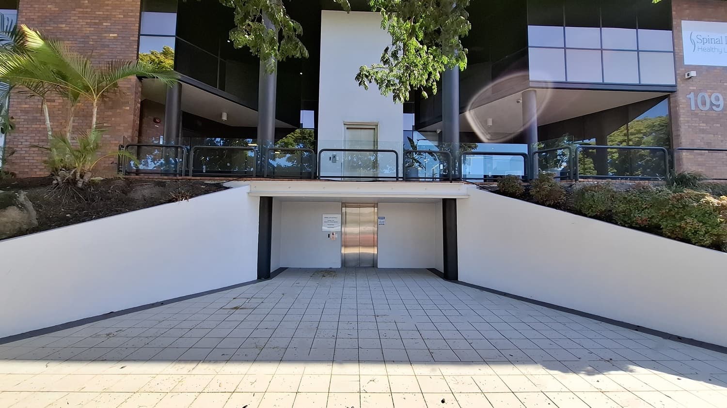 On the left, a brick wall with a small garden area containing palm trees and shrubs. In the middle, a stainless steel lift door set into a white wall, with a tiled ramp leading up to it. On the right, another brick wall with a sign and more greenery. The floor is tiled with light-coloured square tiles, and the ceiling above the lift area is white with recessed lighting.