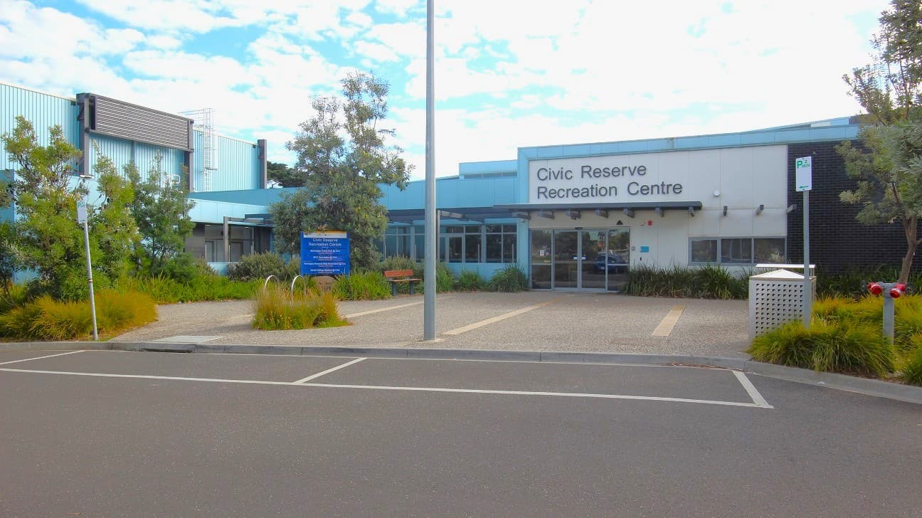 Civic Reserve Recreation Centre is a white and sky blue, low-set building. Bushes and trees surround the venue. Parking car spaces close to the front of the entrance are separated by yellow lines. P 2-minute parking sign in green legend and white background can be seen on both sides of the entryway to car spaces in front of the building. On the left side, near the entrance doors, are a brown wooden bench and a large blue signboard.